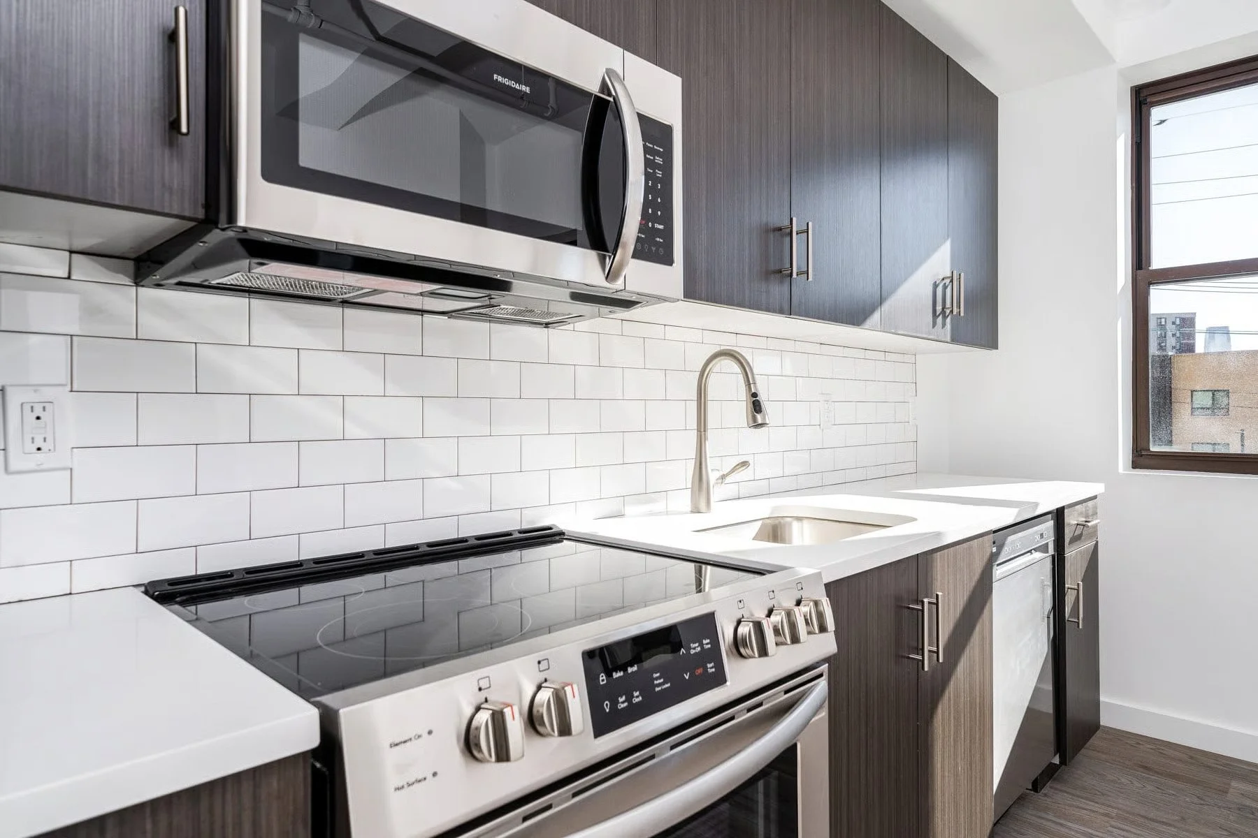 Modern kitchen with white countertop, stainless steel microwave, oven, and small refrigerator, dark wood cabinets, white subway tile backsplash, and a window to the right letting in natural light.