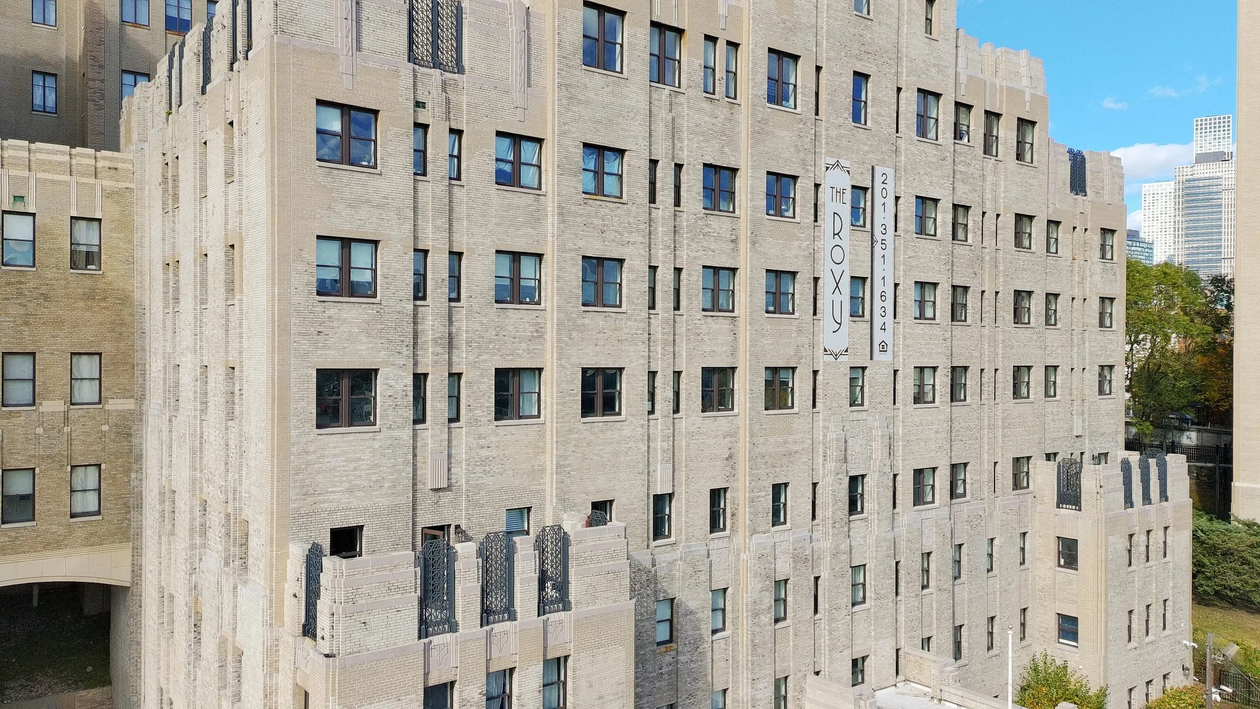A multi-story brick building, The Roxy, with numerous windows and decorative ironwork on the balconies. The background shows a cityscape with high-rise buildings, and the sky is partly cloudy.