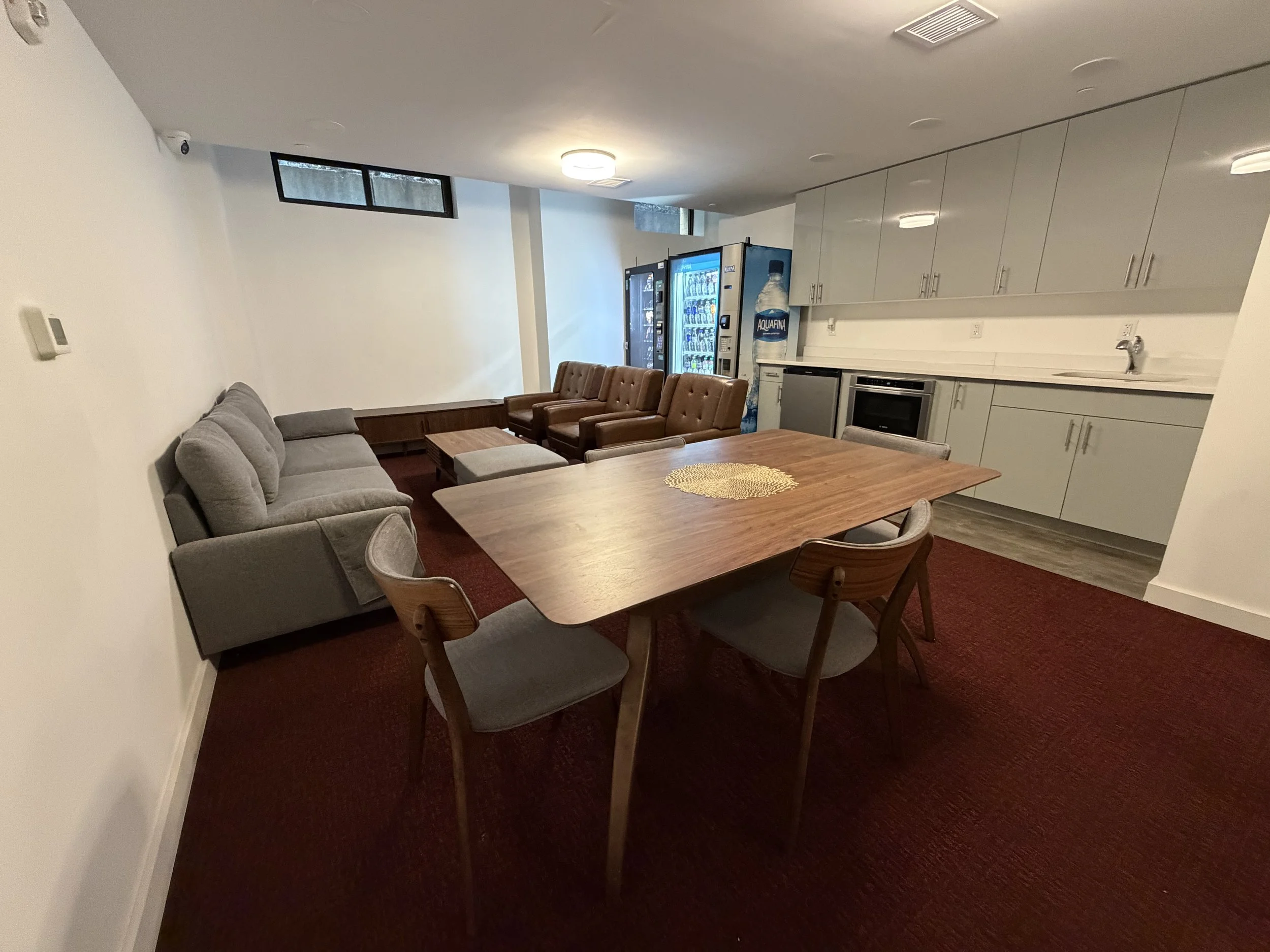 A lounge area with a wooden table, gray chairs, a gray sofa, brown armchairs, a kitchenette with cabinets, and vending machines in the background.
