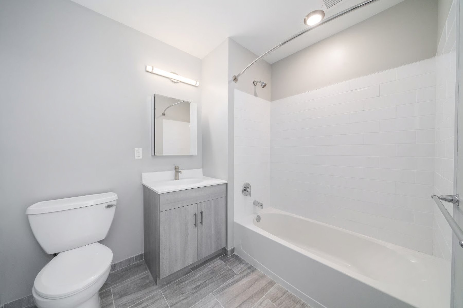 Modern bathroom with a white toilet, a gray vanity with a mirror, and a bathtub with a shower, tile flooring, and gray walls.