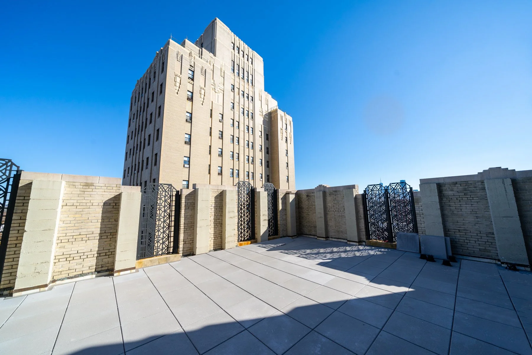 City rooftop terrace with brick and metal fence, concrete flooring, and tall skyscraper against a clear blue sky.