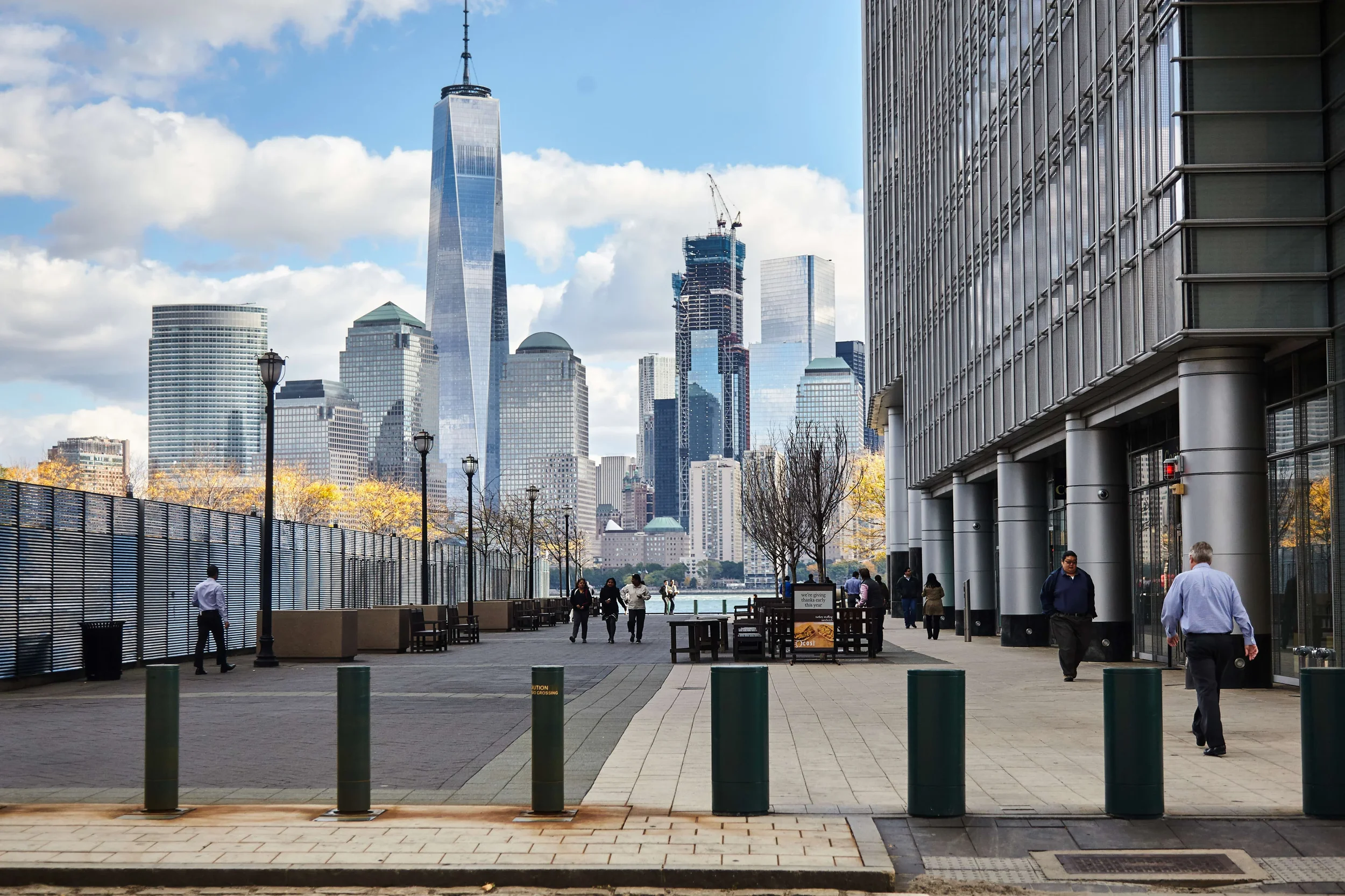 Cityscape with high-rise buildings, including One World Trade Center, along a waterfront walkway with pedestrians, trees, and modern architecture.