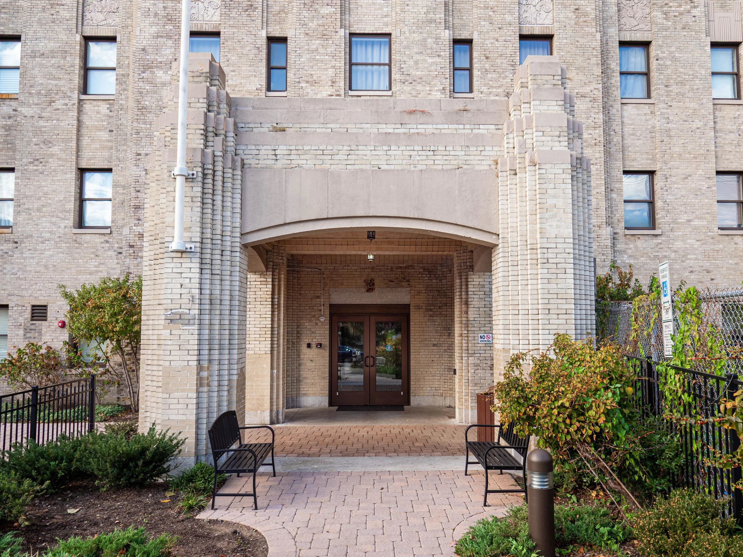 Entrance to a building with brown double doors, brick walls, and arched entryway. There are two black benches outside, surrounded by small landscaped gardens, a lamp post, and parking signs.