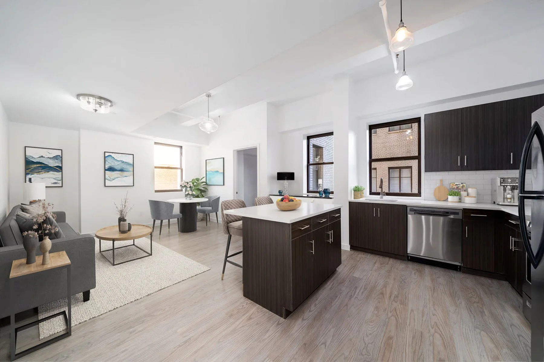 Modern open-concept living room and kitchen with white walls, dark wood cabinetry, stainless steel appliances, and large windows, furnished with gray and neutral-toned furniture and decor.