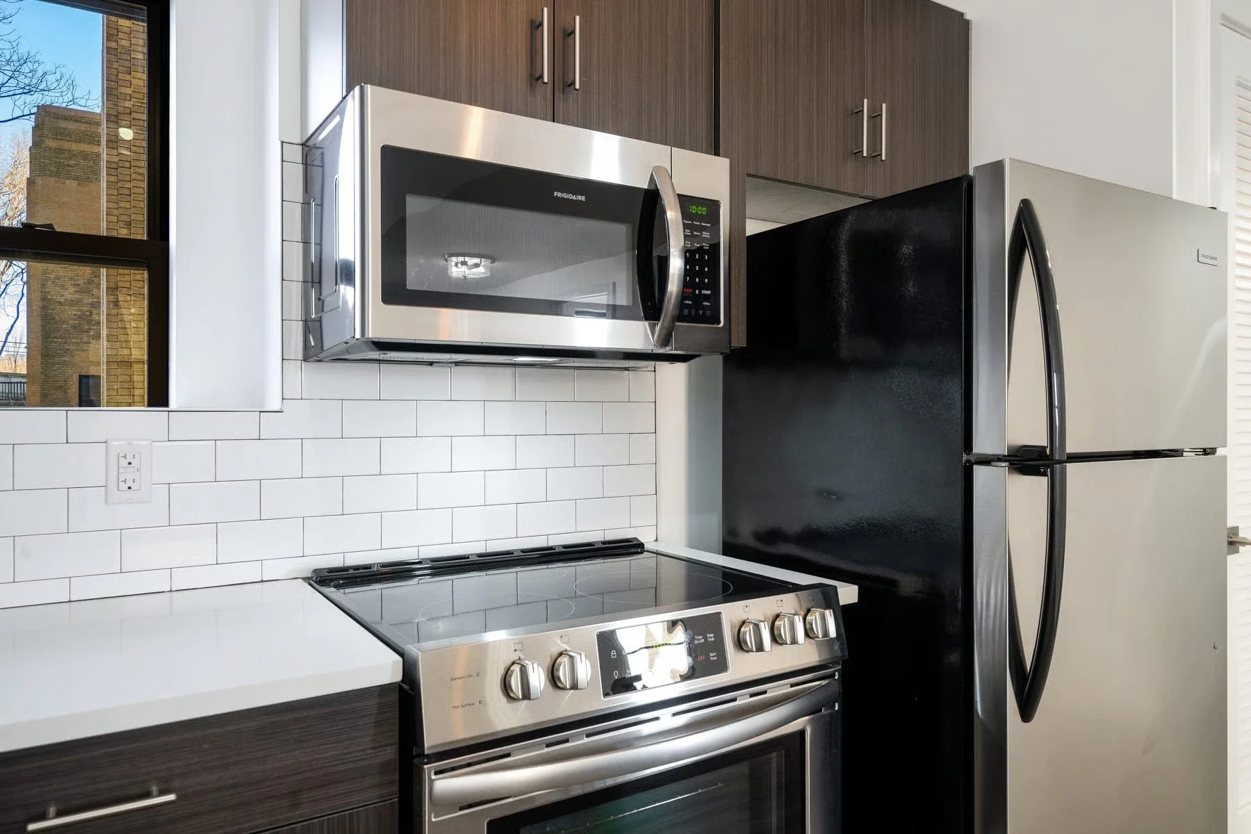 Modern kitchen with white subway tile backsplash, stainless steel appliances including a microwave, oven, and refrigerator, and dark brown cabinets.