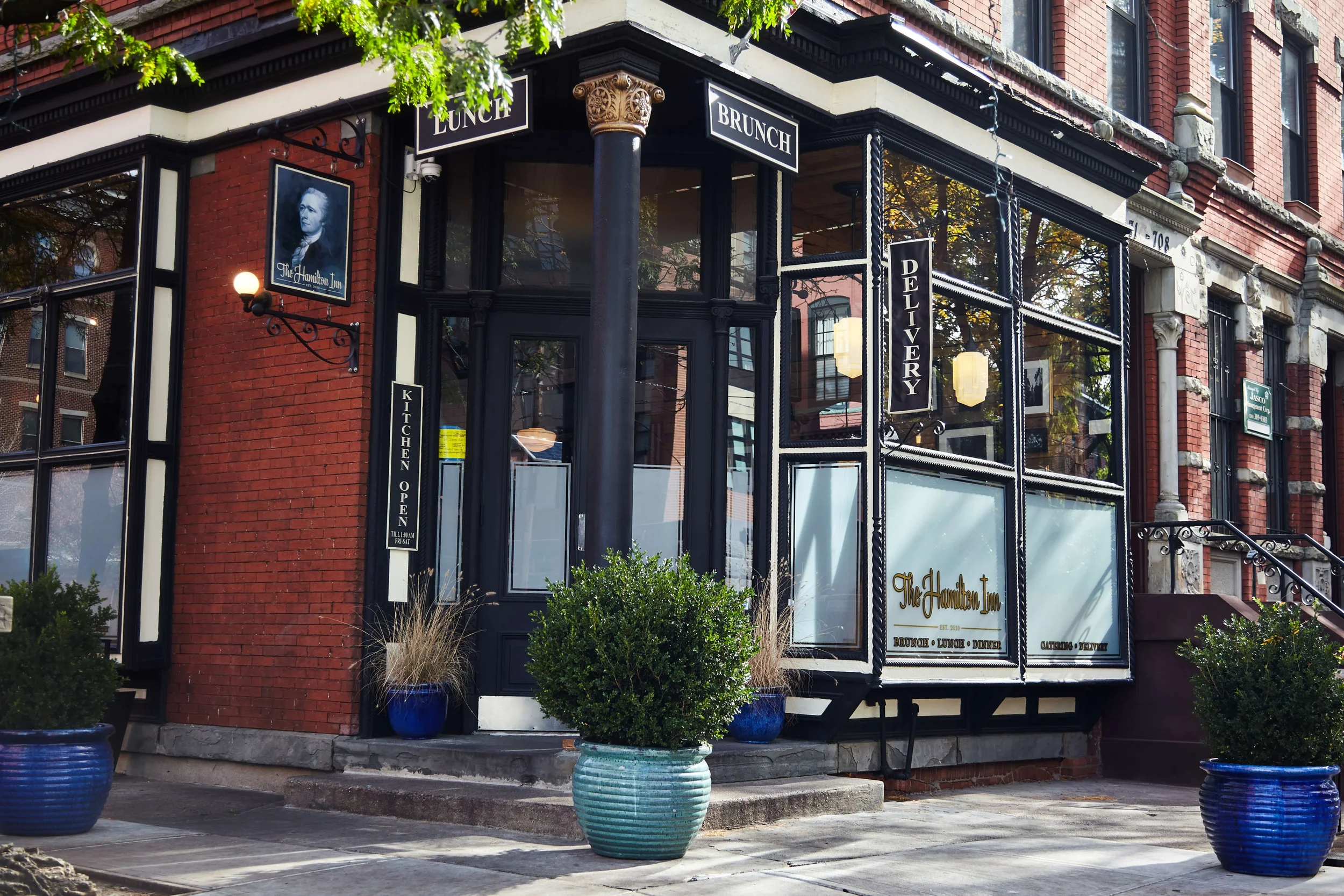 Exterior of a restaurant with brick and black-painted facade, large windows, potted plants outside, and signs indicating it's open for brunch, lunch, and dinner.