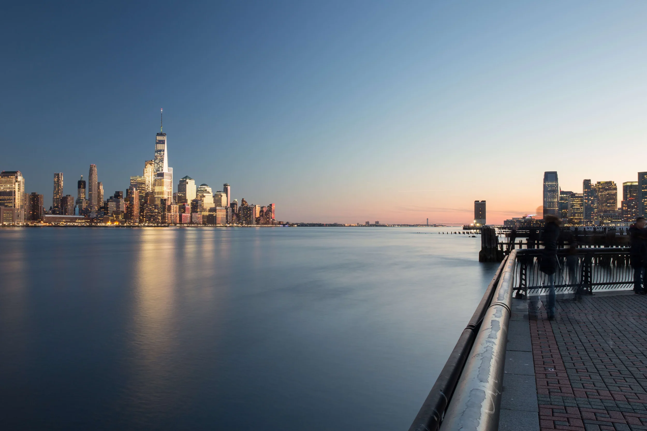 View of the Manhattan skyline at dusk from a waterfront promenade, with calm water reflecting city lights and some people standing by the railing.