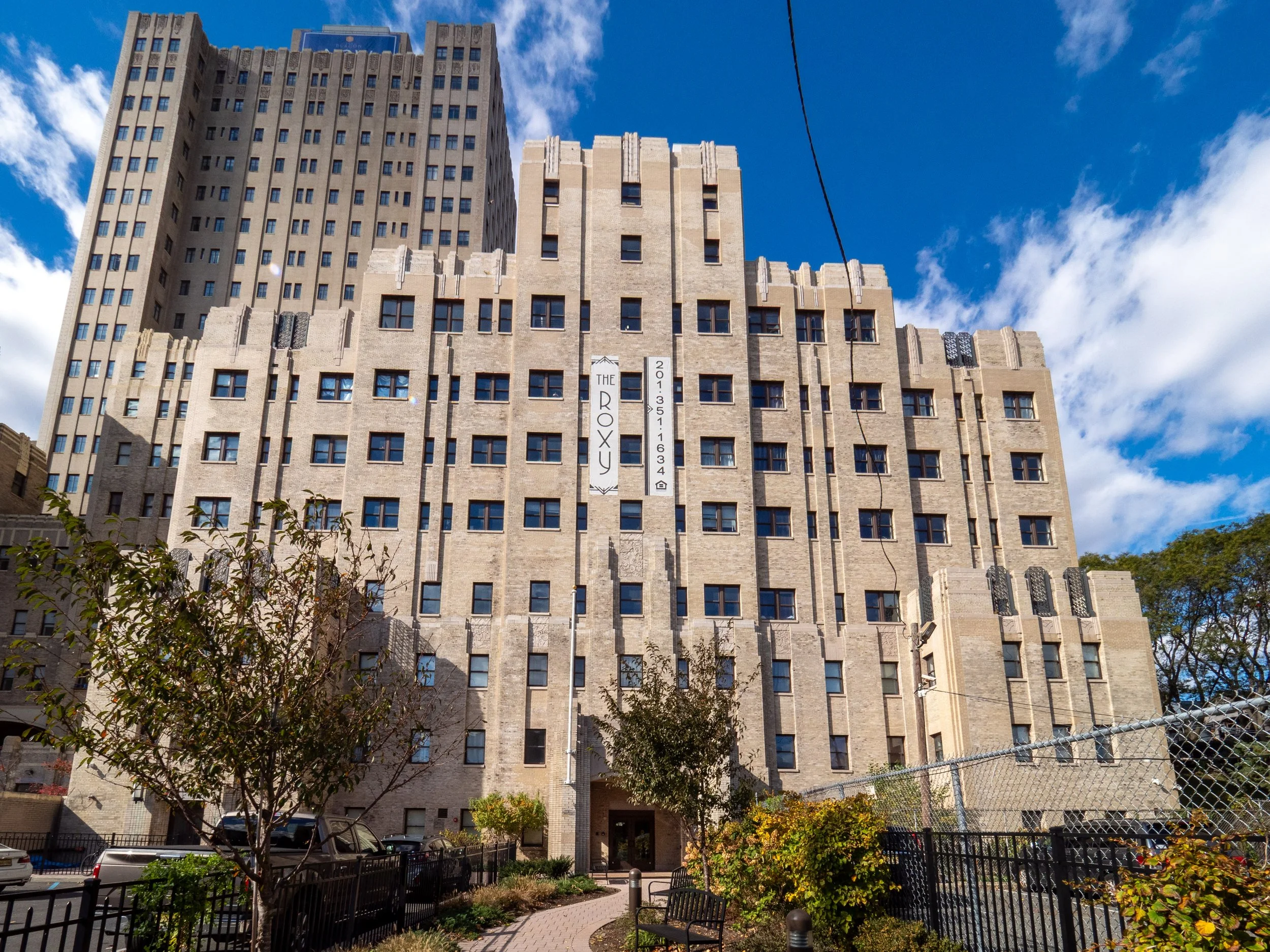 A tall beige brick apartment building has multiple windows, some with black decorative elements, and is set against a blue sky with some clouds. There are trees, a small garden, and a pathway with benches in the foreground, and cars parked nearby. 