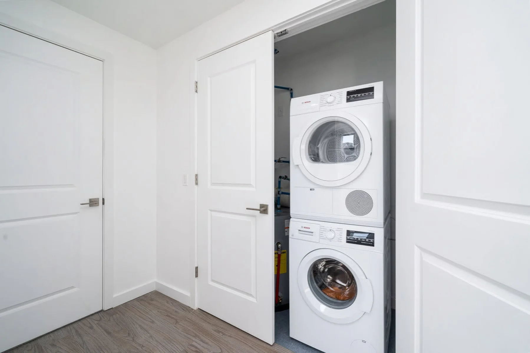A laundry closet with a stacked washer and dryer, partially open double doors, white walls, and wood floor.
