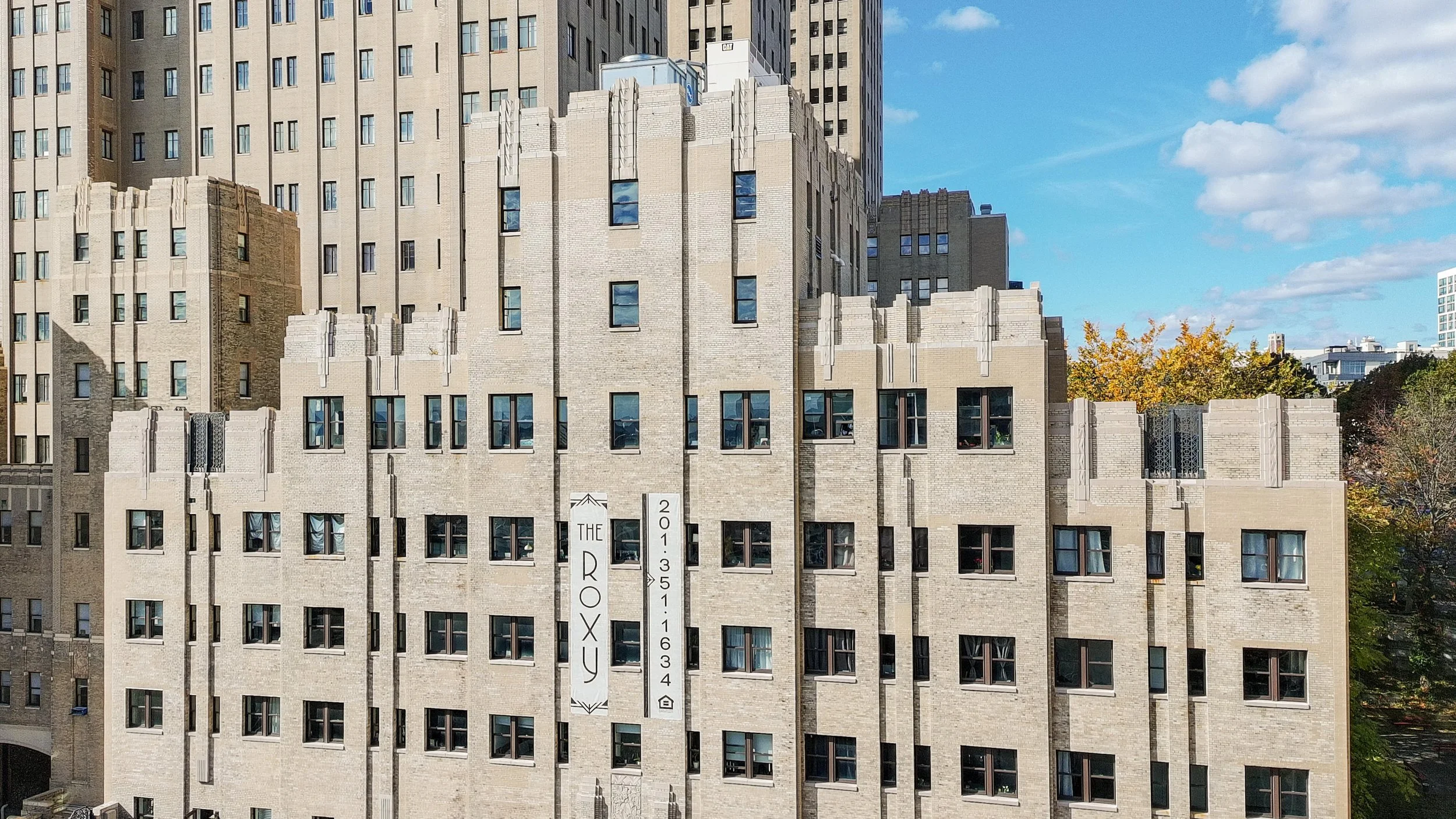 Facade of a multi-story building named The Roxy with a stone exterior, several windows, and a sign with trees and a blue sky in the background.