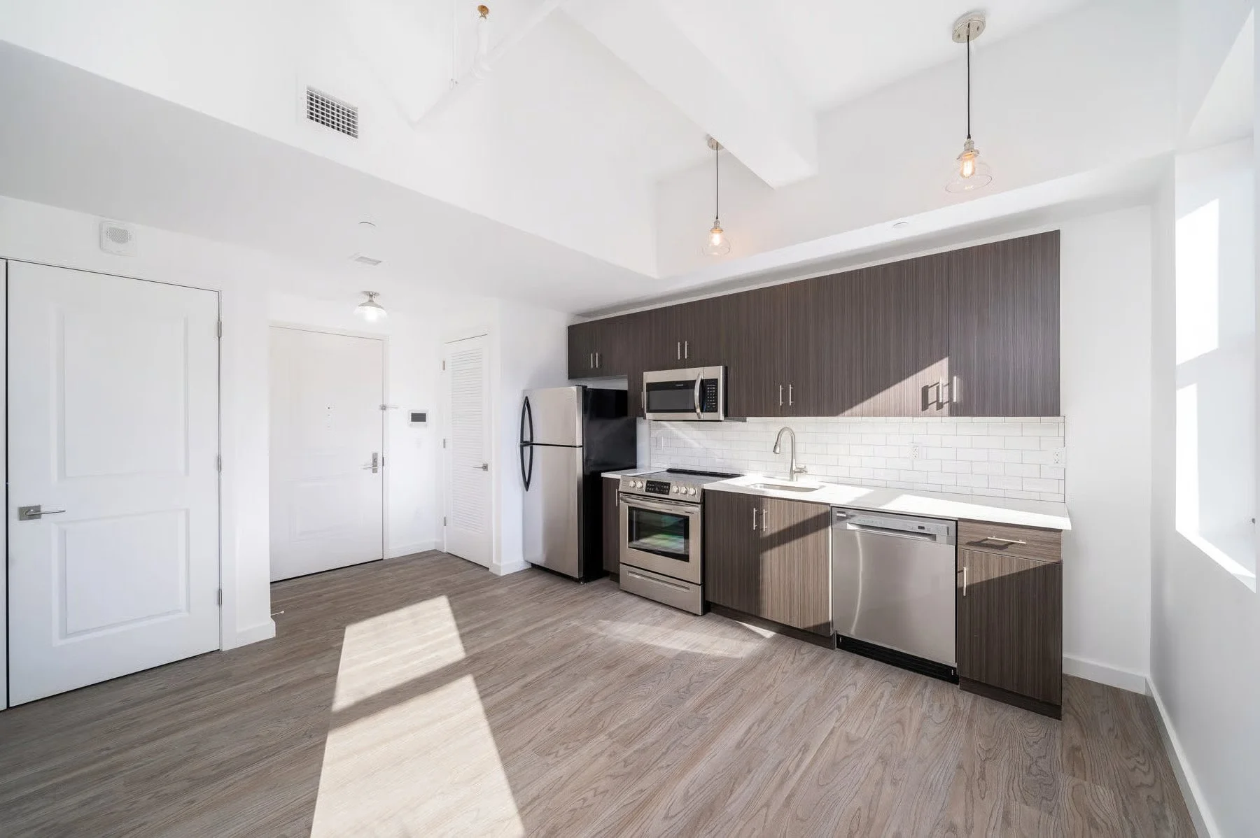 Modern kitchen with white walls, wood cabinets, stainless steel appliances, white subway tile backsplash, and light wood flooring, illuminated by natural light and hanging light fixtures.