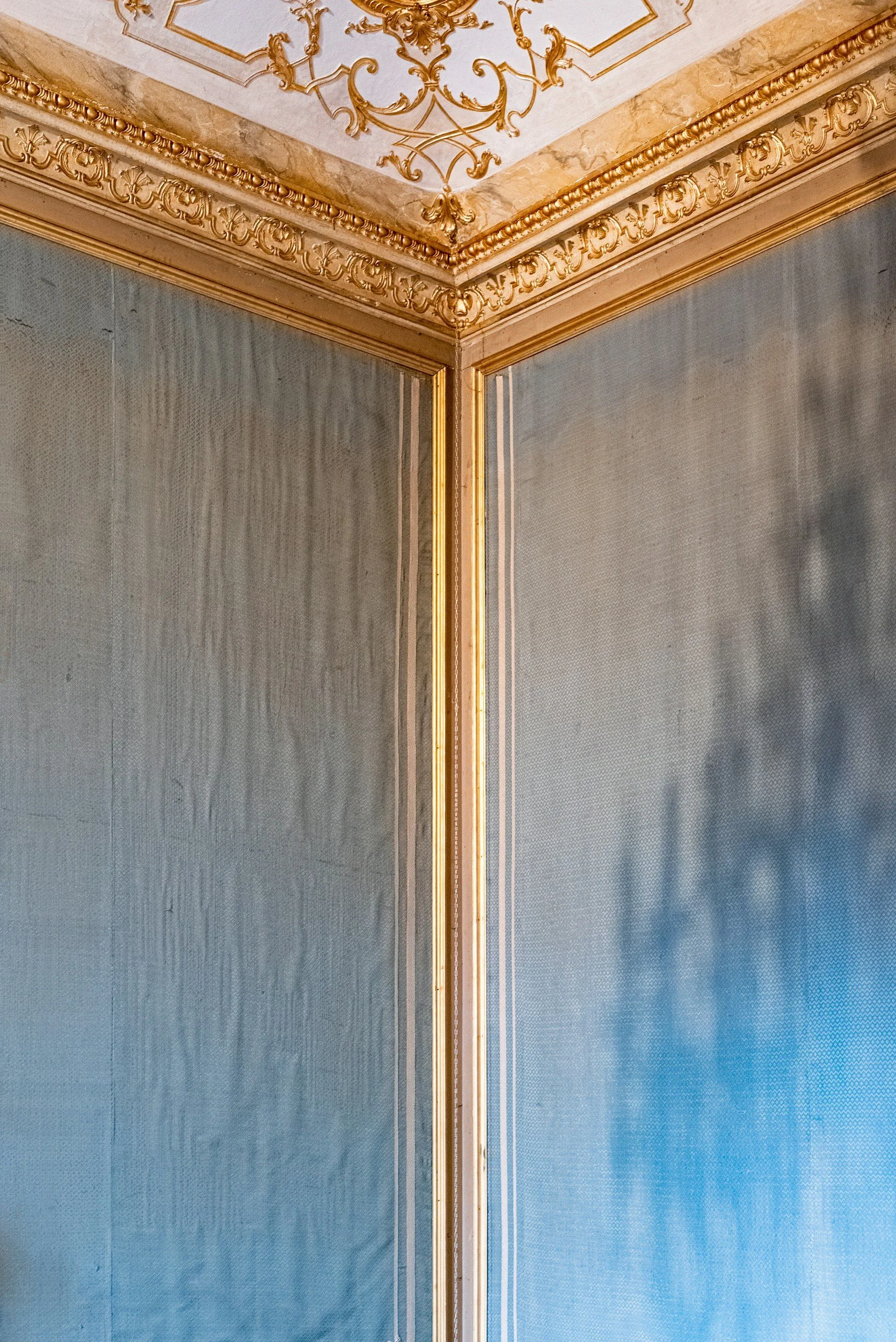 Close-up of an ornate corner of a room with gold decorative moldings, detailed ceiling embellishments, and textured blue walls.