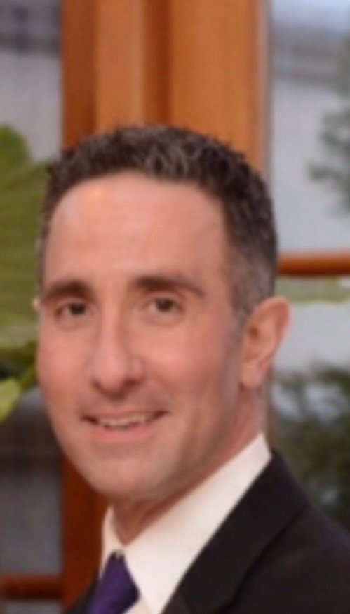 A middle-aged man in a suit and tie smiling at the camera, indoors with a wooden window frame and outside view in the background.