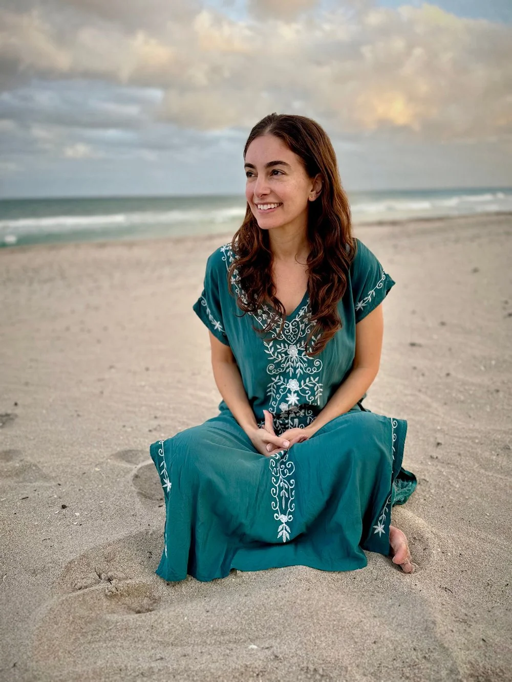 Brooke Novick, wearing a teal embroidered dress, is sitting cross-legged on a sandy beach, smiling, with the ocean and cloudy sky in the background.