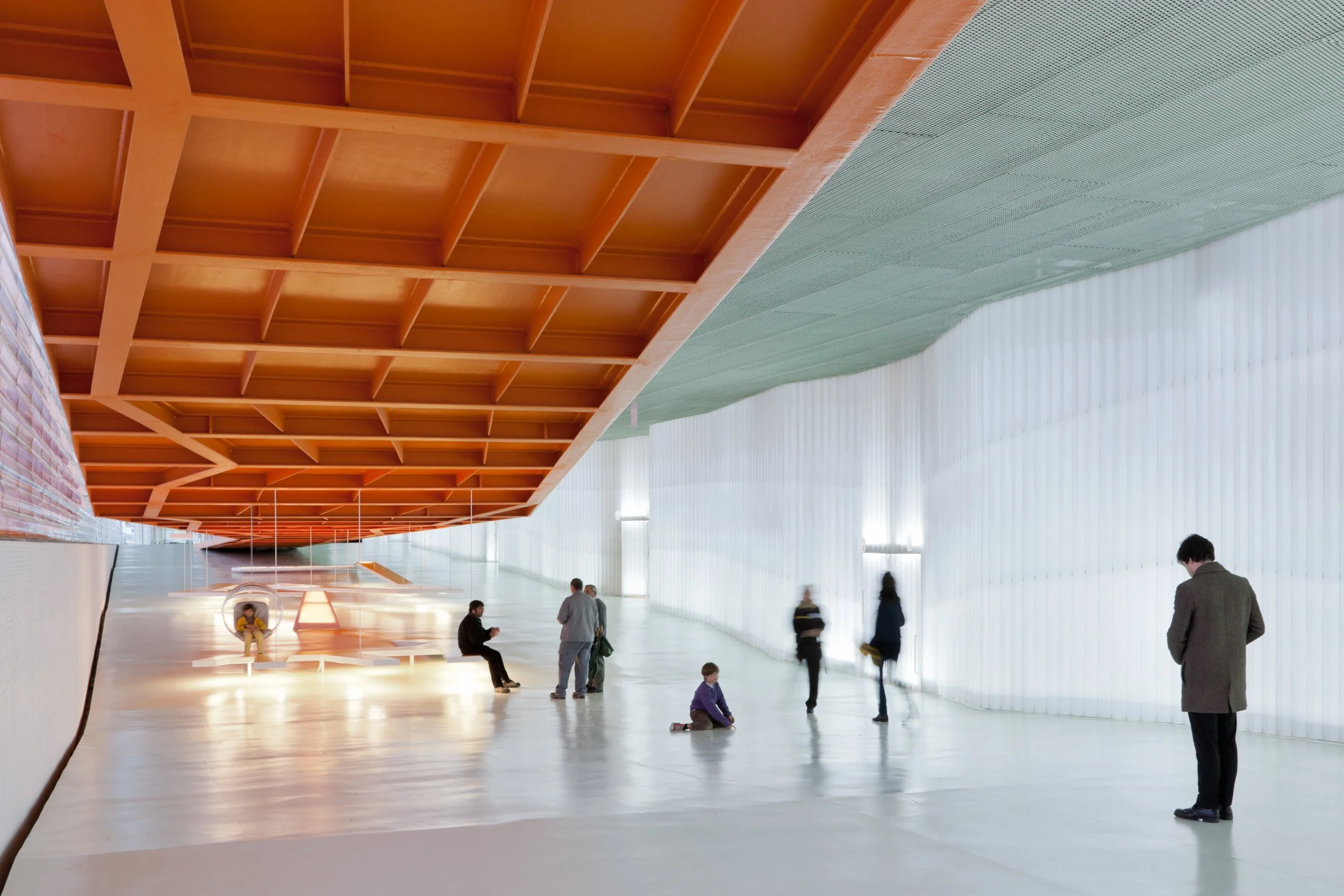 Interior de un edificio con techo de madera y paredes blancas, donde varios adultos y niños están en un espacio amplio y luminoso, algunos sentados y otros de pie, con iluminación suave y ambiente moderno.