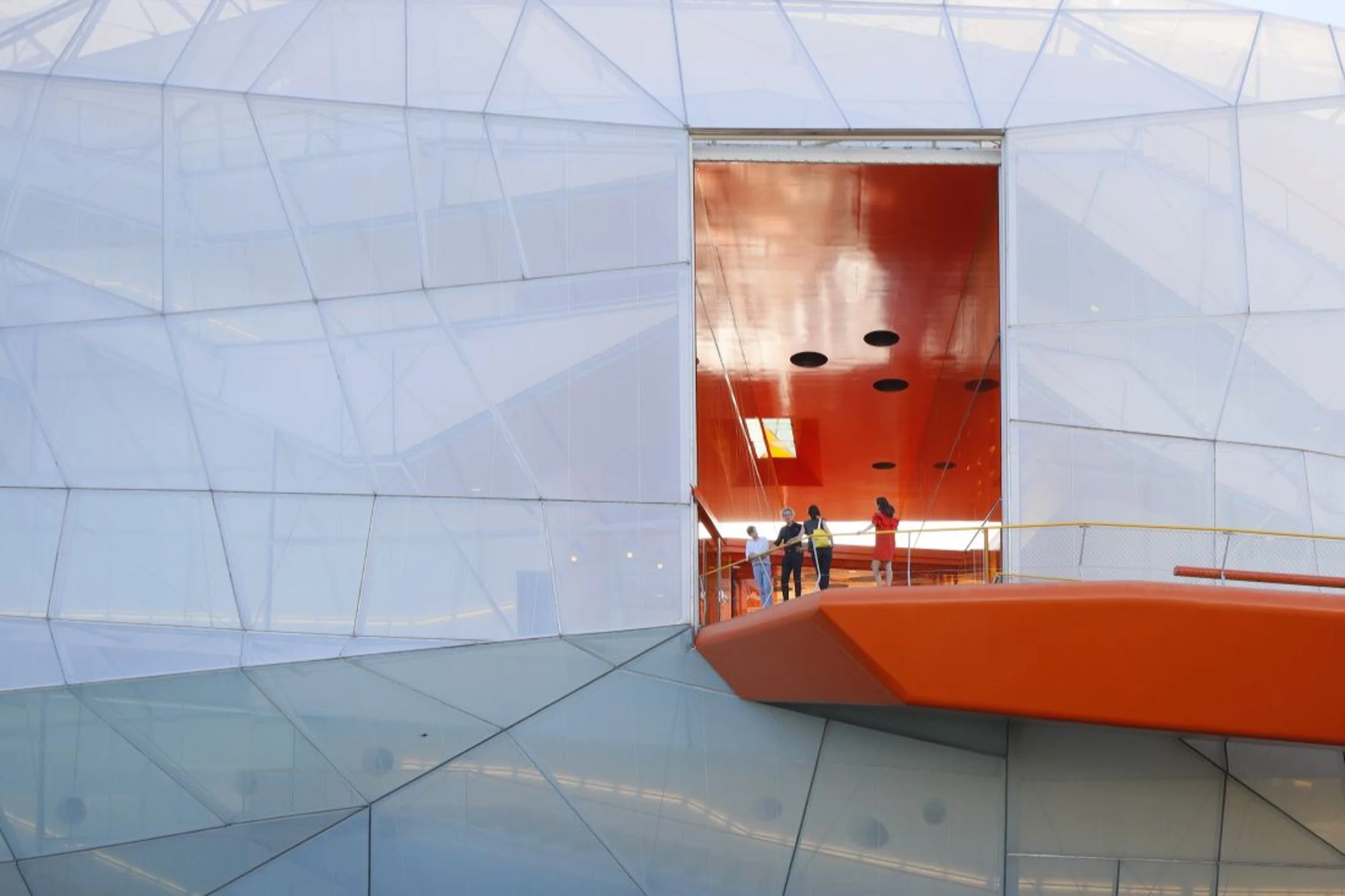 Interior de un edificio moderno con paredes de paneles de cristal y un techo en forma de cúpula. Se observan varias personas en una plataforma con barandillas naranjas explorando el espacio.