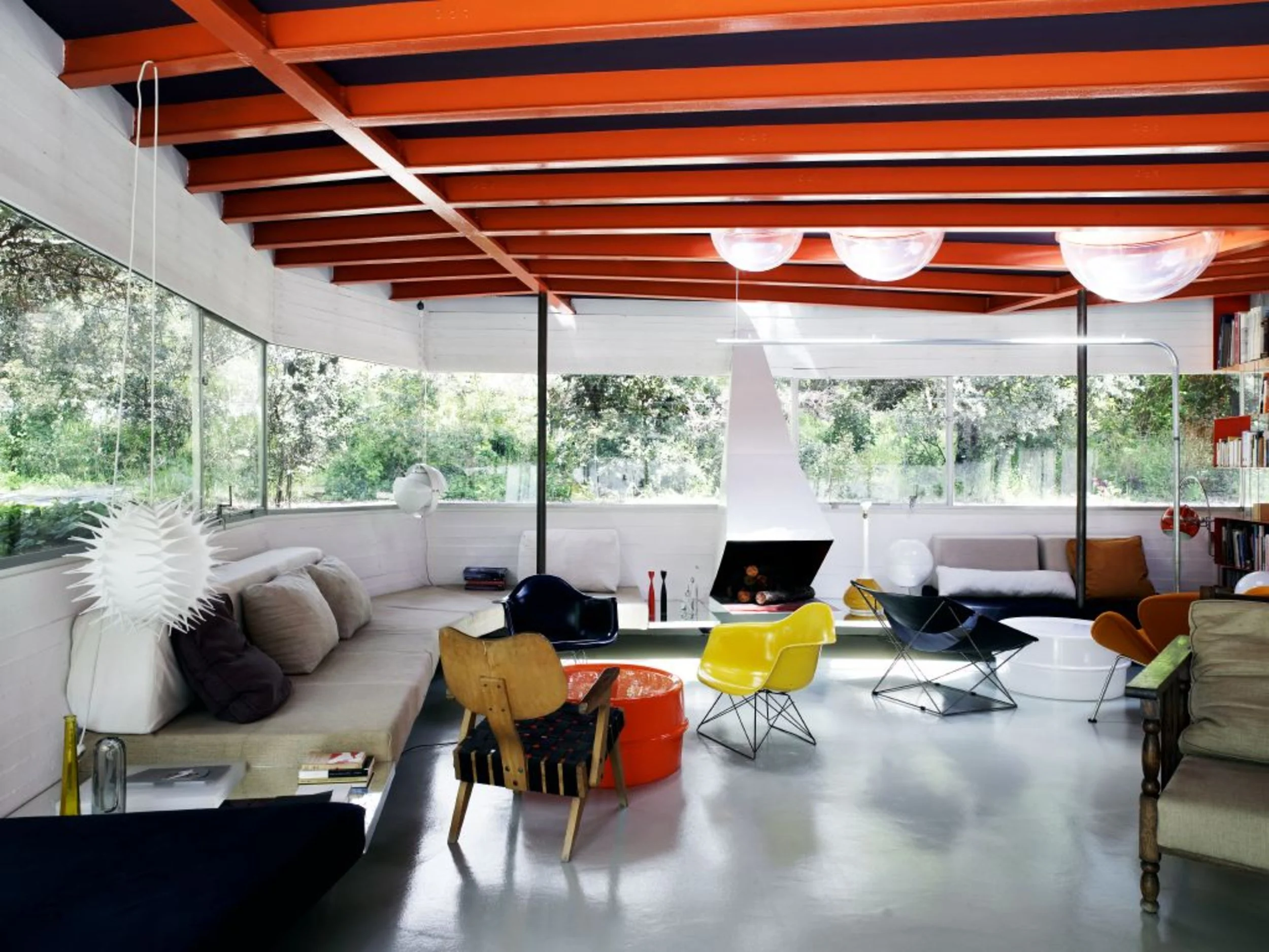 Interior de una sala moderna con muebles coloridos, grandes ventanas con vista a la naturaleza, iluminación natural y techo de madera con vigas rojas.