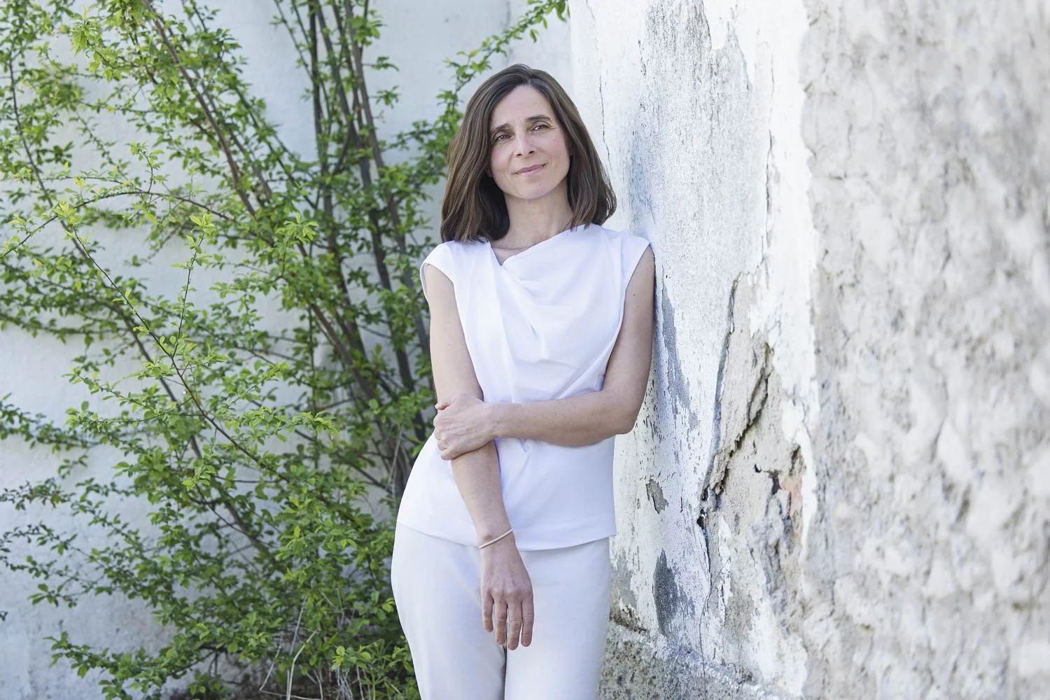 Mujer con vestido blanco, apoyada en una pared de piedra, con plantas verdes de fondo, sonrisa suave.
