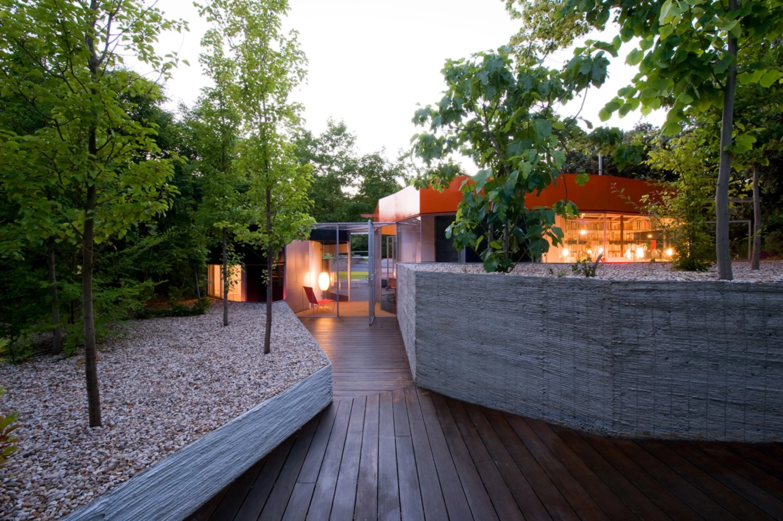 Casa moderna rodeada de árboles y vegetación, con área de descanso iluminada en la noche, piso de madera y paredes de concreto en un entorno natural.