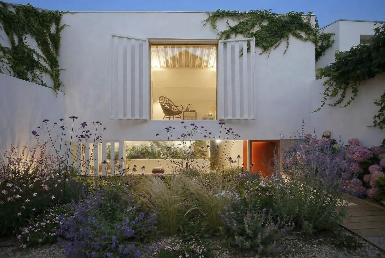 Casa moderna de dos pisos con paredes blancas, ventanas con cortinas, jardín con plantas y flores, y un interior iluminado visible desde afuera.