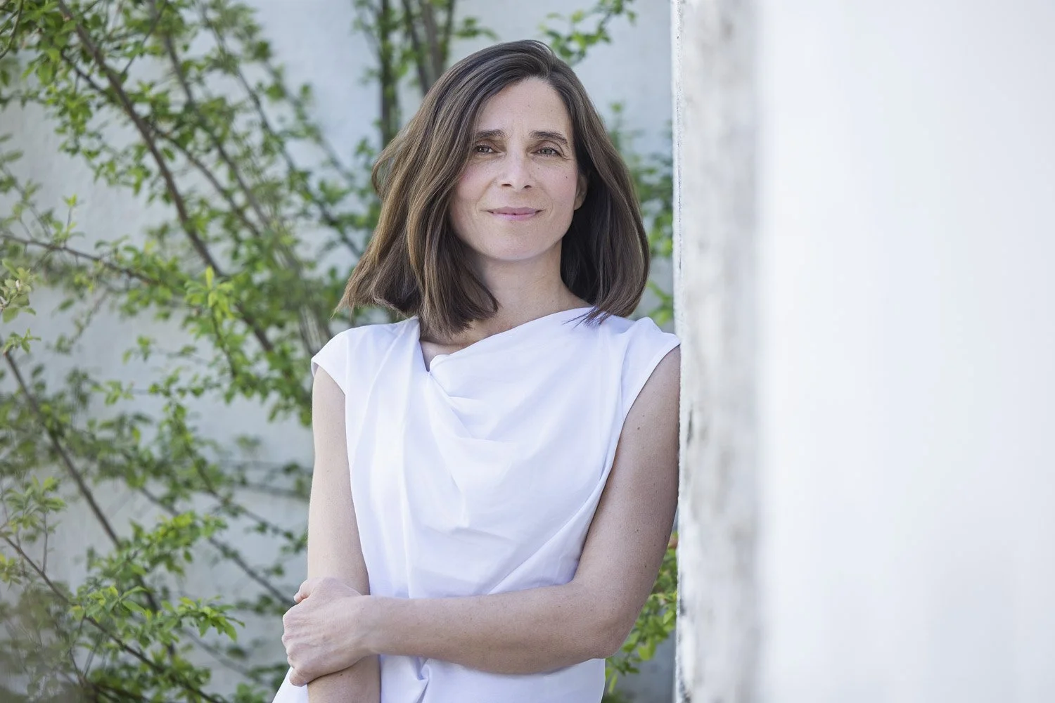 Mujer con cabello castaño oscuro, usando blusa blanca, de pie junto a una pared y vegetación de fondo.
