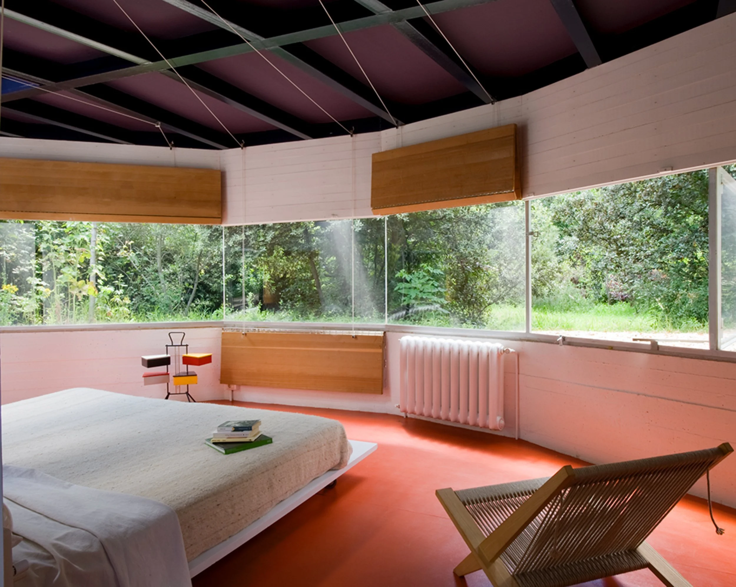 Interiores de una casa con grandes ventanas de vidrio que permiten ver un jardín con árboles, muebles de madera y paredes blancas con radiador a la derecha.
