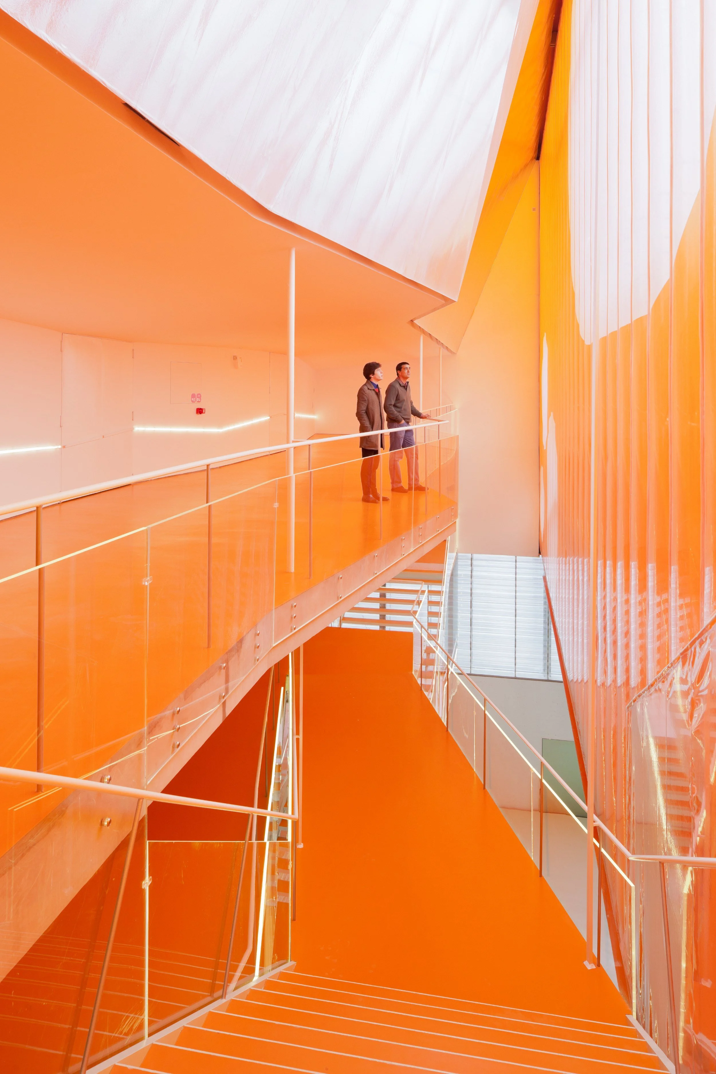 Vista interior de un edificio con paredes y escaleras pintadas en color naranja brillante, y dos personas de pie en una pasarela observando hacia el interior.