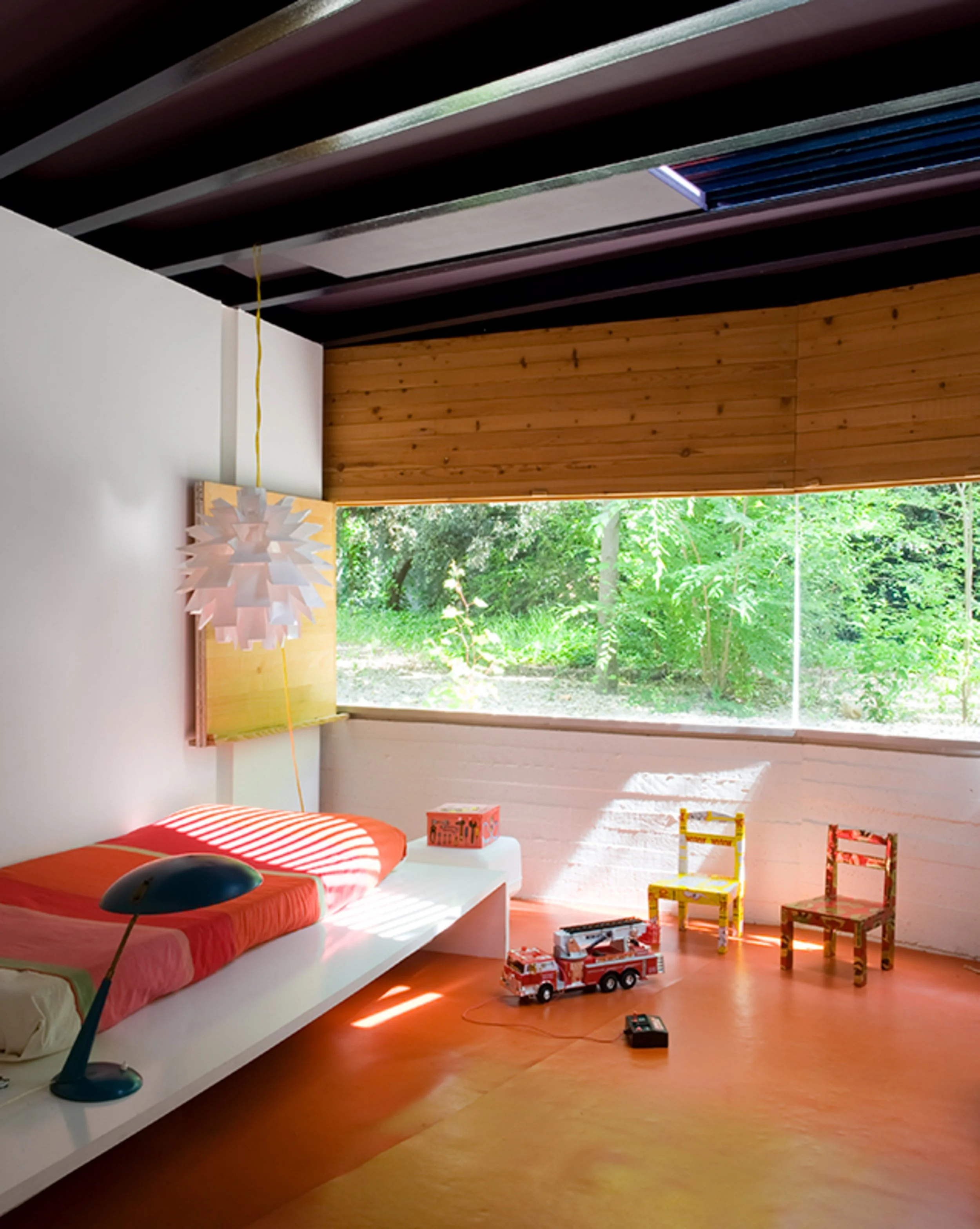 Habitación infantil con cama, juguetes, sillas, ventana con vista a naturaleza, lámpara de diseño, piso de madera y paredes blancas y de madera.