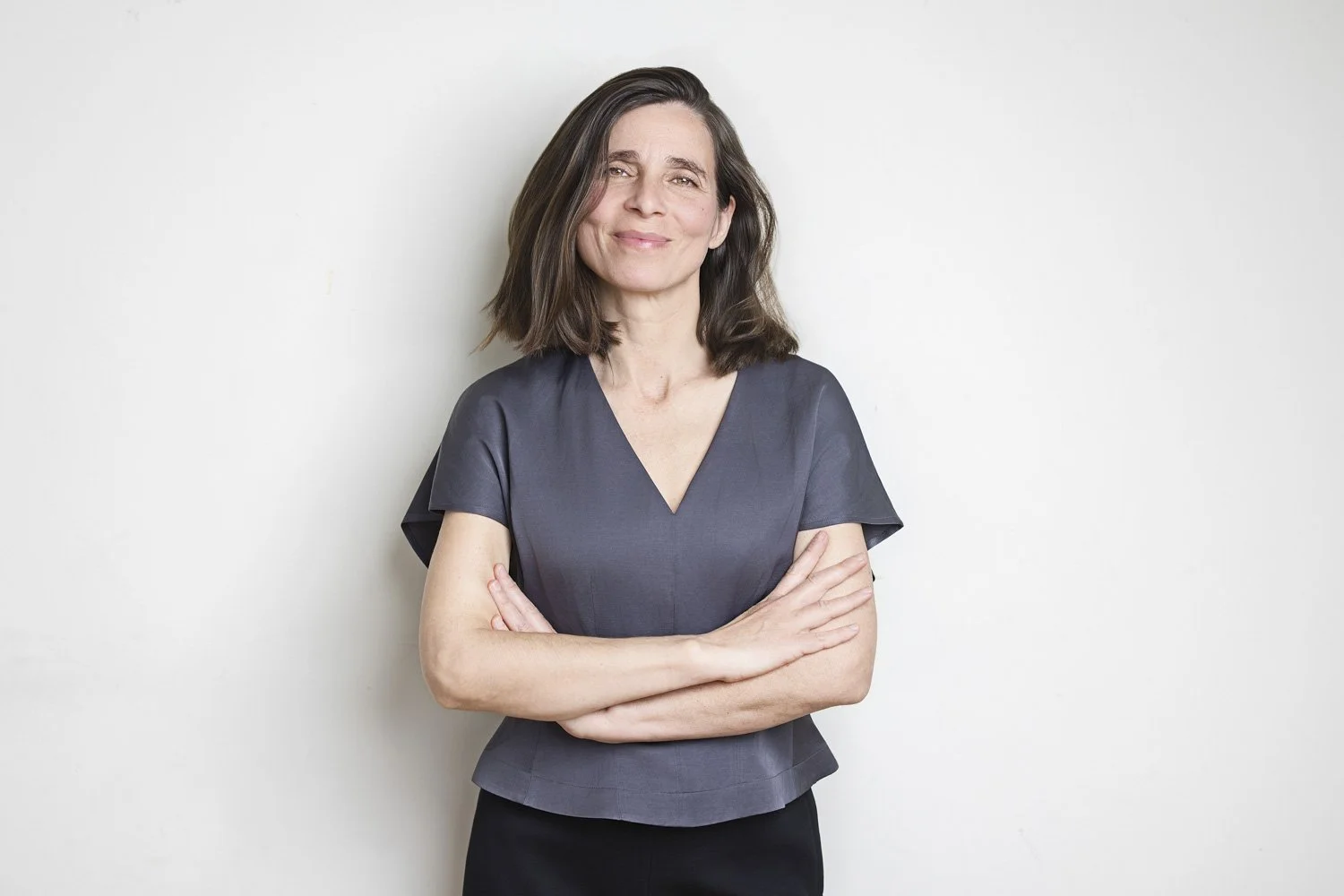 Lara Resco, vistiendo blusa gris y pantalón negro, de pie contra fondo blanco.