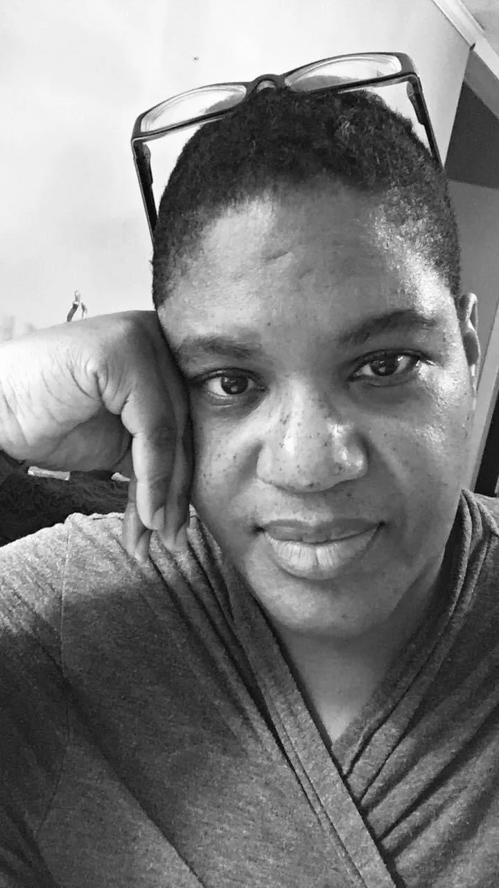 Close-up black and white photo of a person with short hair, resting their face on their hand, wearing glasses on their head, and smiling slightly.