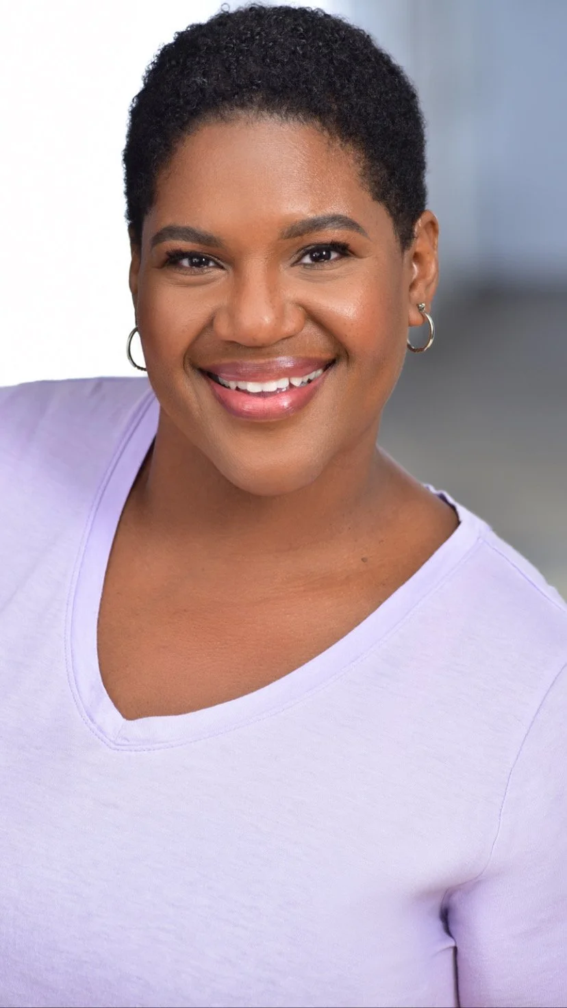 Portrait of a smiling Black woman with short curly hair, wearing hoop earrings and a light purple shirt, against a blurred background.