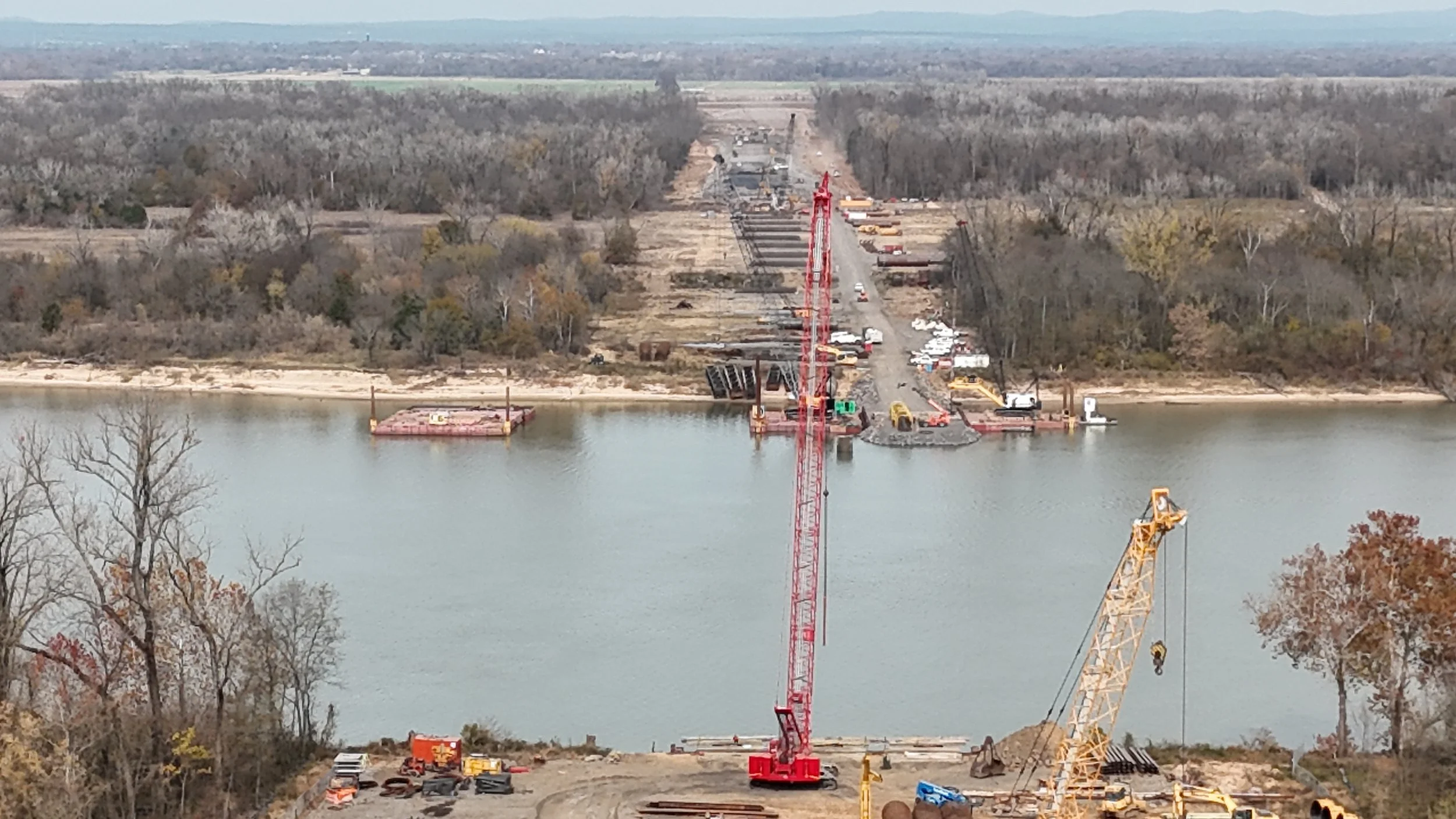 Construction site across a body of water with cranes and heavy machinery building a bridge or structure, surrounded by trees and distant landscape.