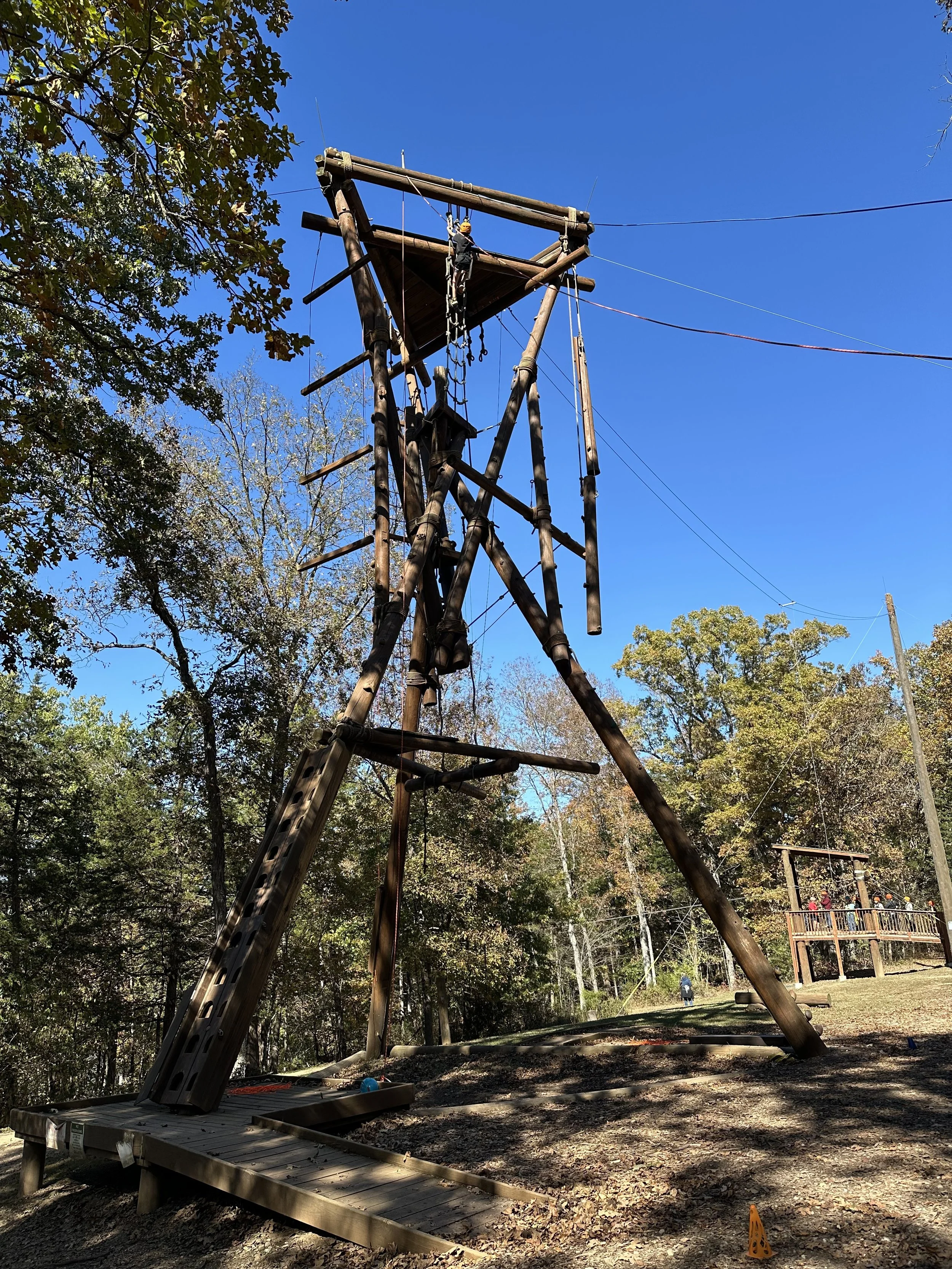 A tall wooden adventure ropes course tower with climbing elements, situated outdoors in a wooded area with a clear blue sky.