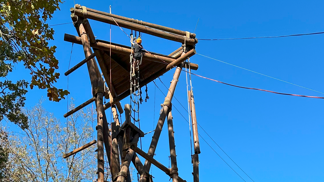 A person wearing a helmet climbing an outdoor wooden ropes course against a bright blue sky, with trees in the background.