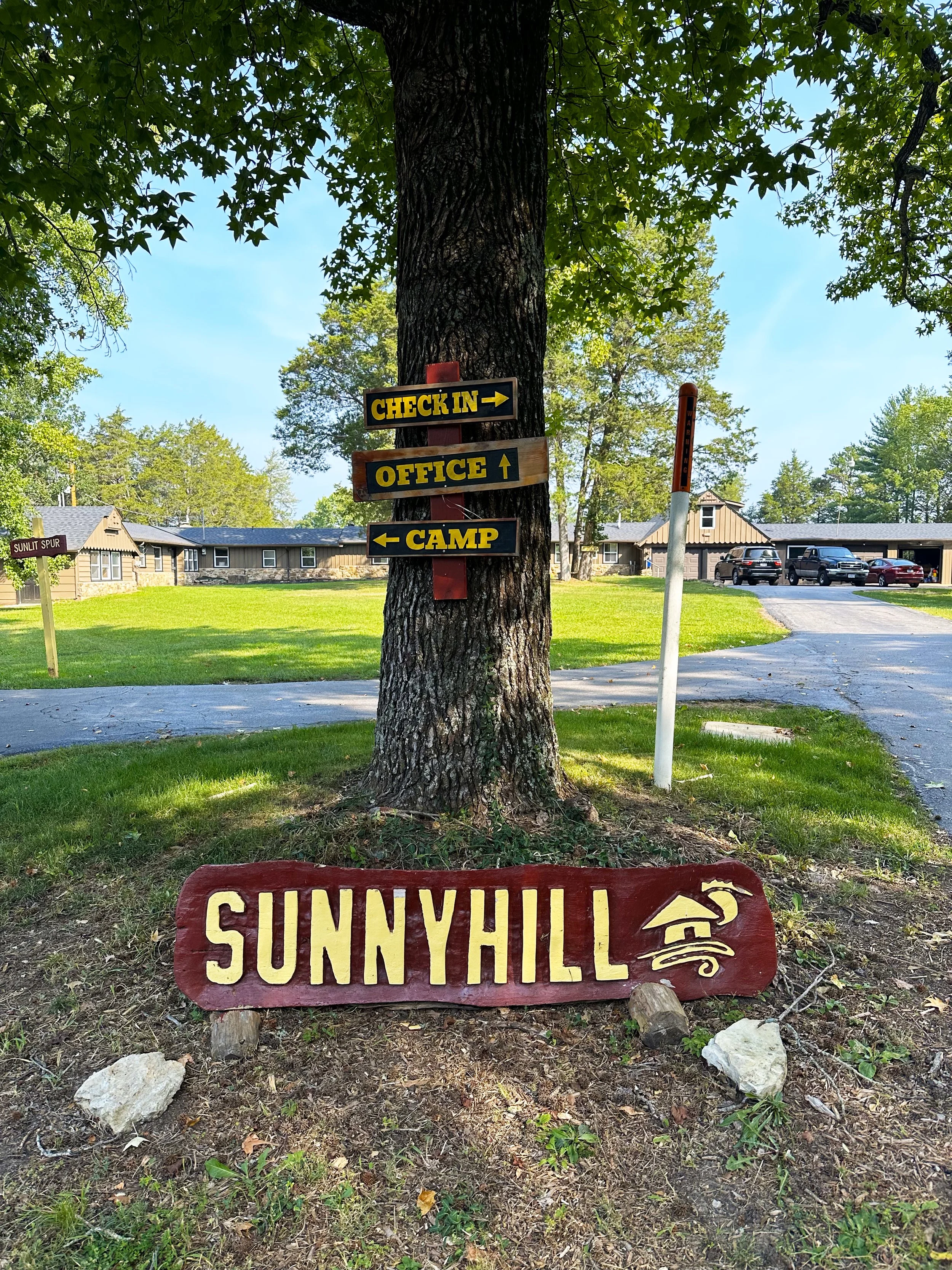 Signage on a tree in a campground area with directions to check-in, office, and camp, and a large wooden sign reading "Sunnyhill" with a camper icon.
