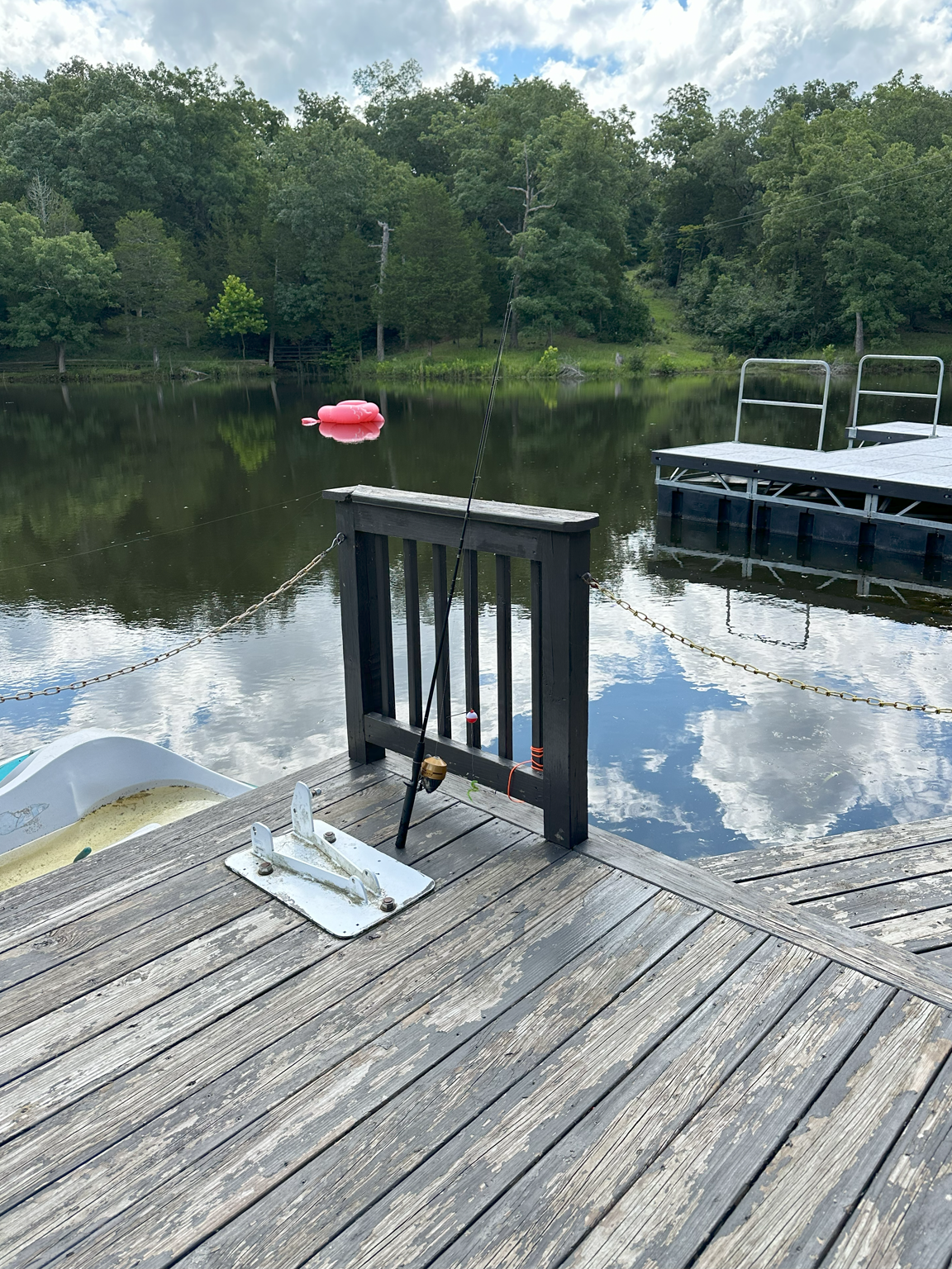 A fishing rod resting on a wooden dock overlooking a calm lake with a pink float, surrounded by green trees under a cloudy sky.