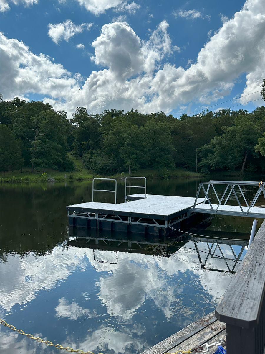 Dock with metal railing on a calm lake, surrounded by green trees under a partly cloudy sky.
