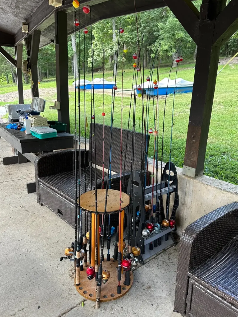 Multiple fishing rods with colorful reels organized in a rack under a outdoor shelter, with a grassy backyard and a small pool in the background.