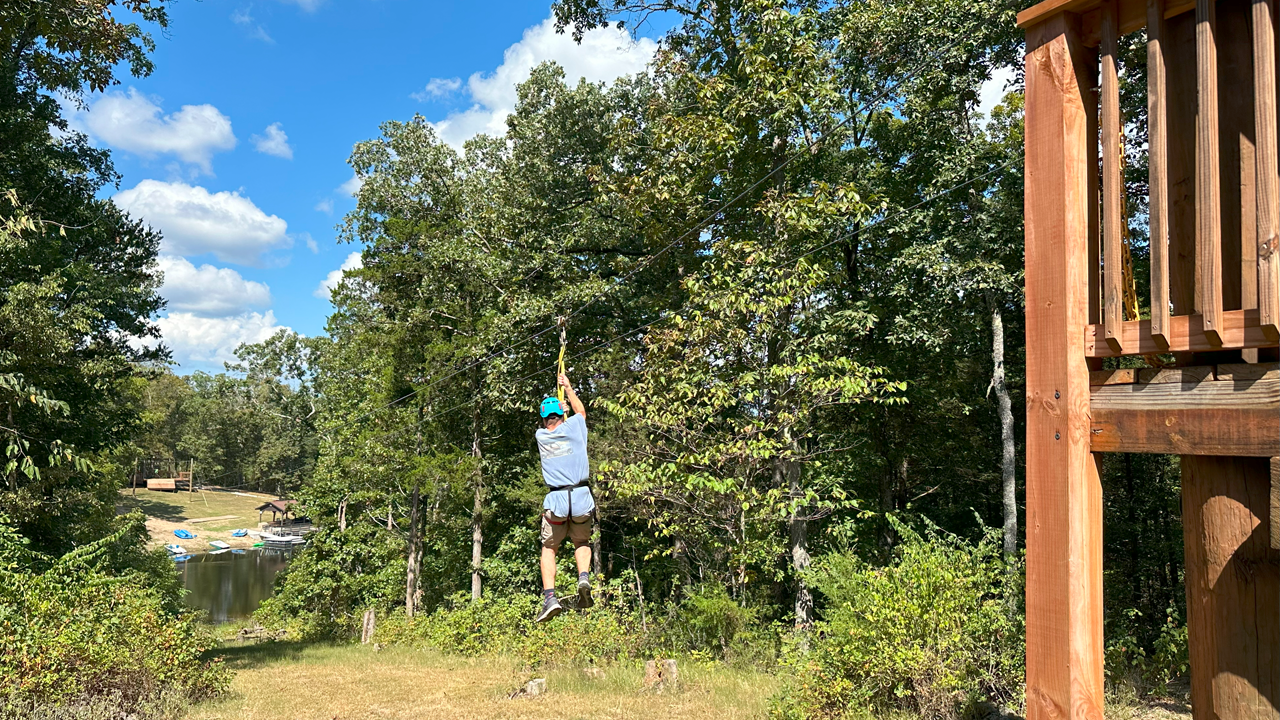 Person wearing a helmet and harness on a zip line in a forested area with tall trees and a blue sky.