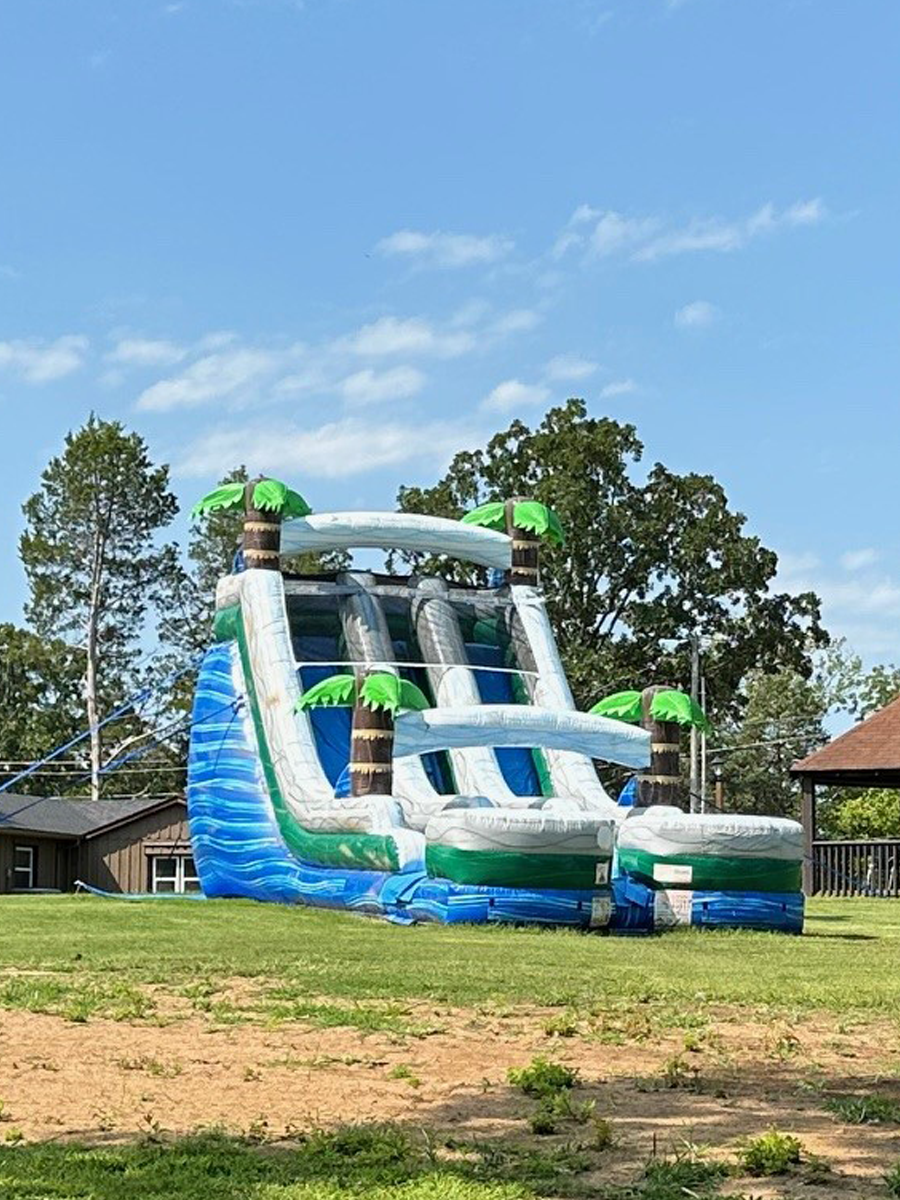 Inflatable outdoor water slide designed to look like a tropical island with palm trees and waterfalls, set on grassy area with trees and houses in background.