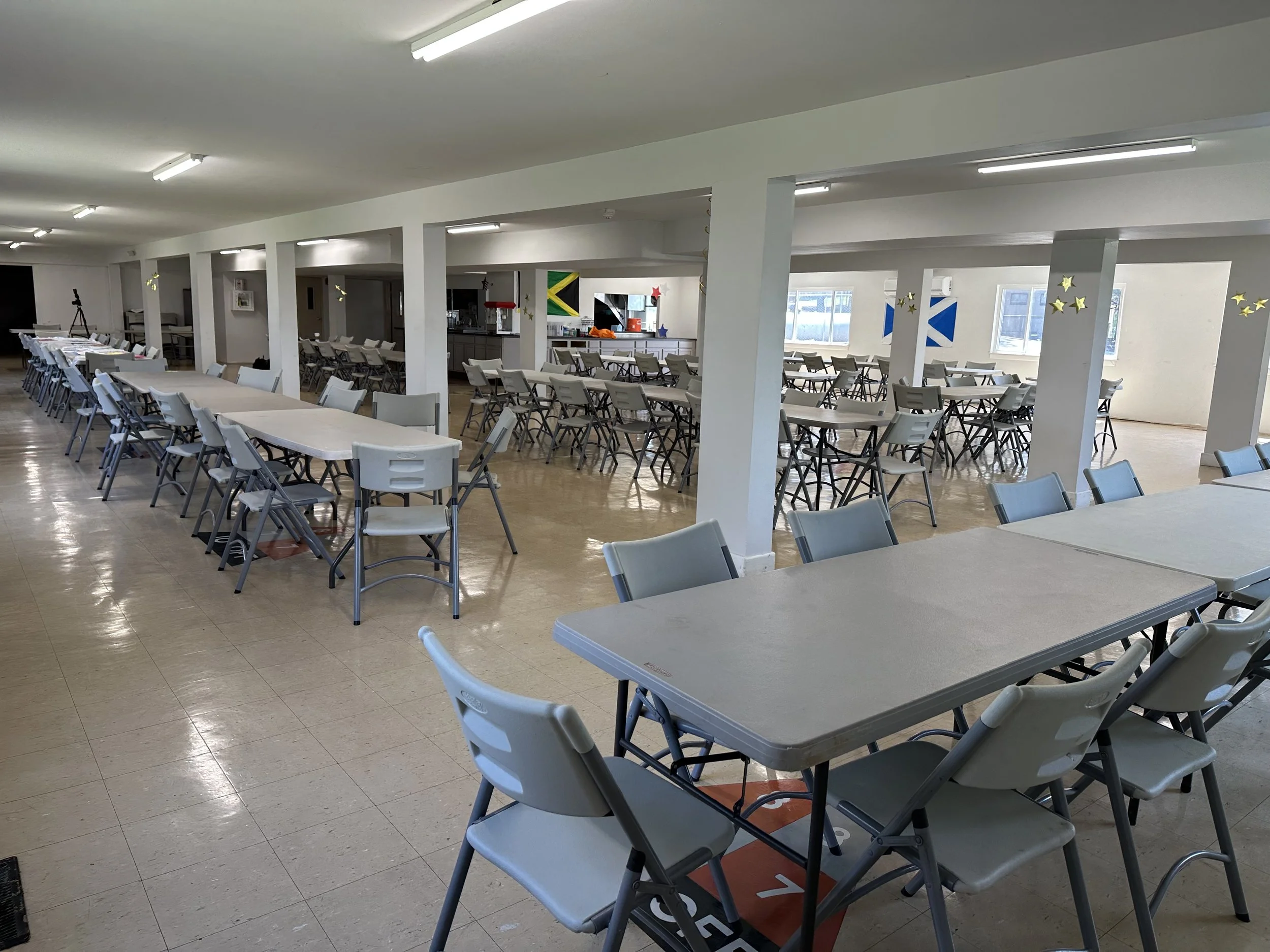 Empty large cafeteria with multiple tables and chairs, some tables covered with white tablecloths, decorated with stars and flags, large windows allowing natural light.