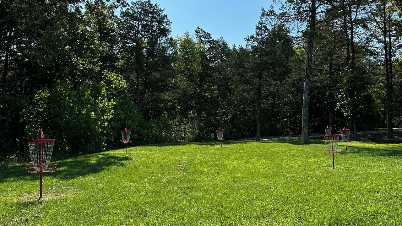 A grassy field with six red and white disc golf baskets surrounded by trees and under a blue sky.