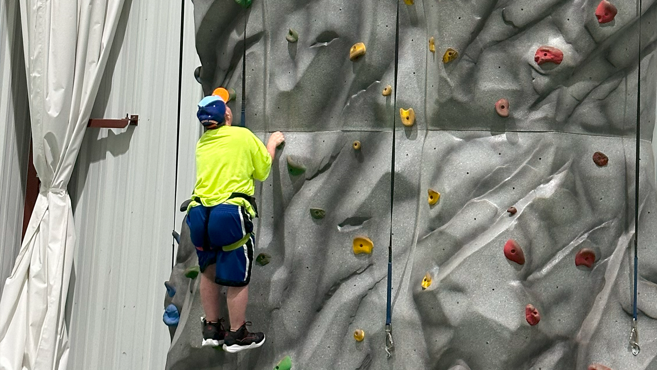 A person in a neon yellow shirt and blue shorts climbing an indoor rock climbing wall with colorful holds.