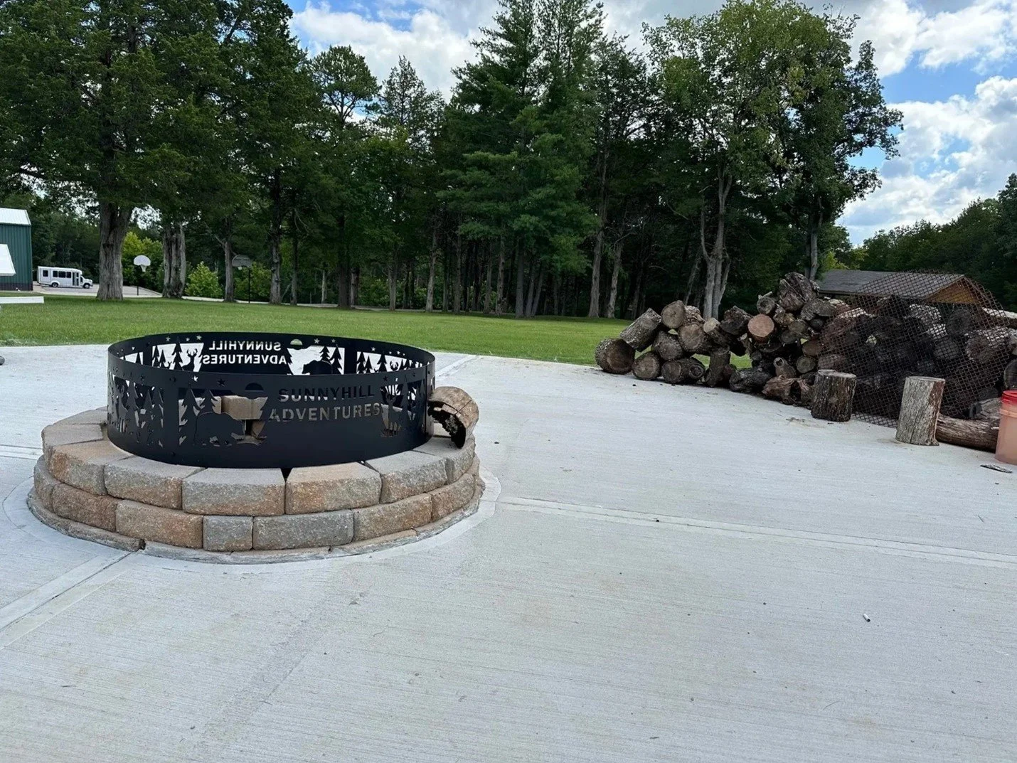 Outdoor scene with a metal fire pit on a brick circle, surrounded by a concrete patio, with stacked firewood on the right, green grass, trees, and a blue sky with clouds in the background.