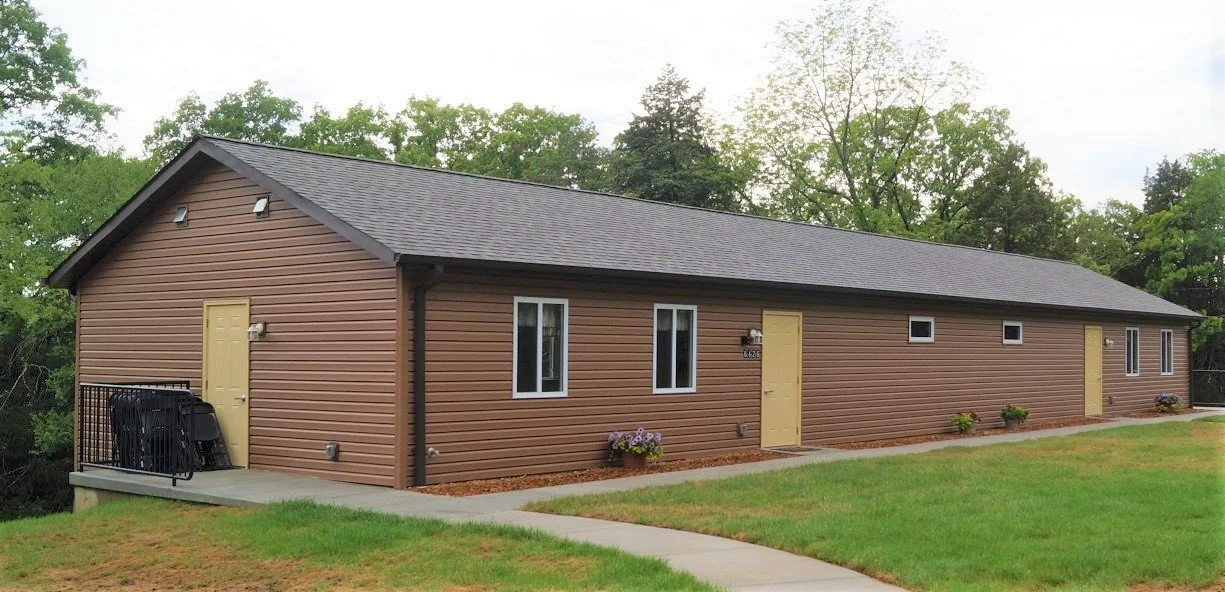 A long, brown, single-story residential building with yellow doors and white-framed windows, situated on a grassy lot with a sidewalk. The roof is dark gray, and there are small potted plants and a patio area with black railings visible on the left side.