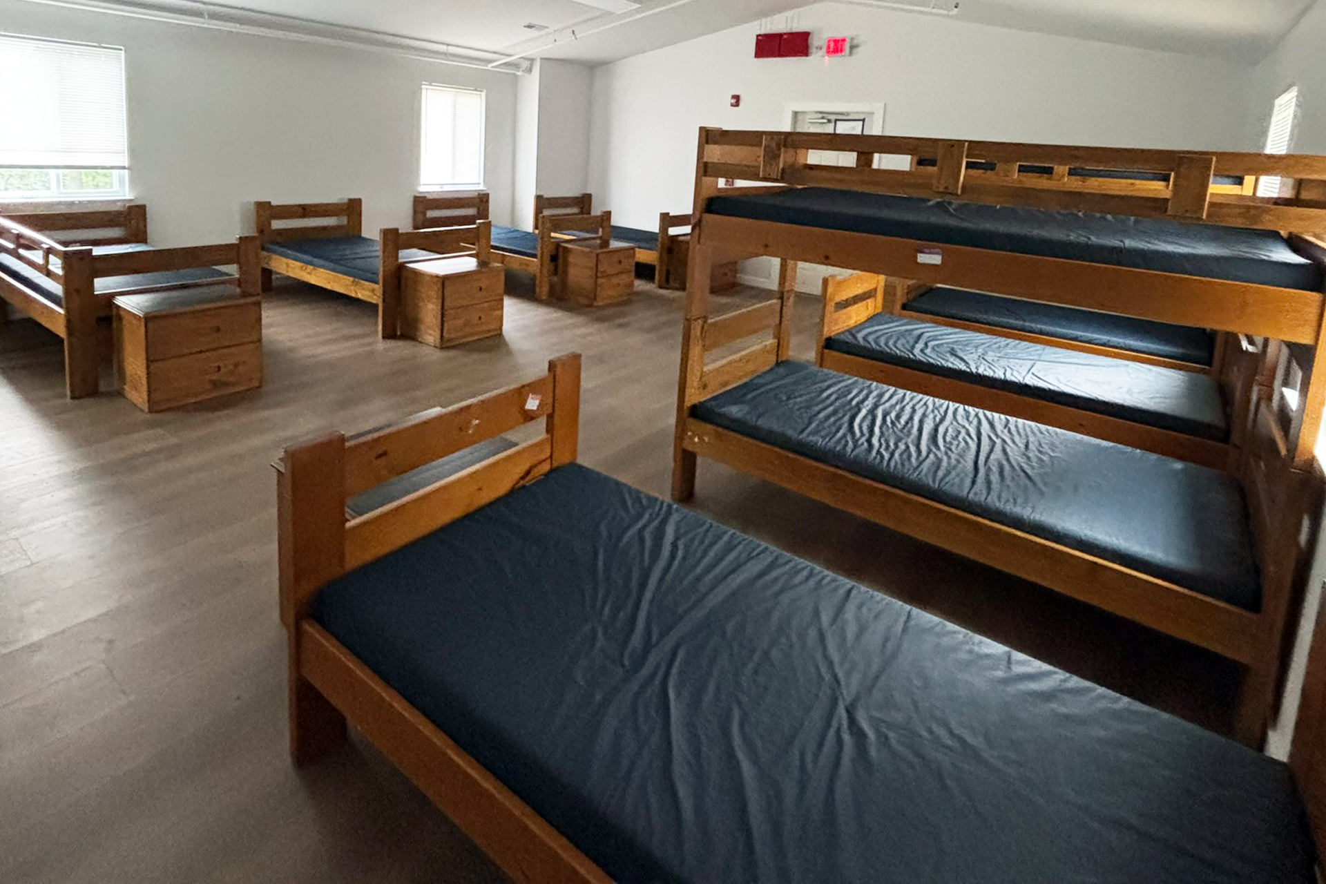 Room with multiple wooden bunk beds with black mattresses, arranged in two rows, with small wooden nightstands next to some beds, windows with blinds on the walls, and a door at the back.