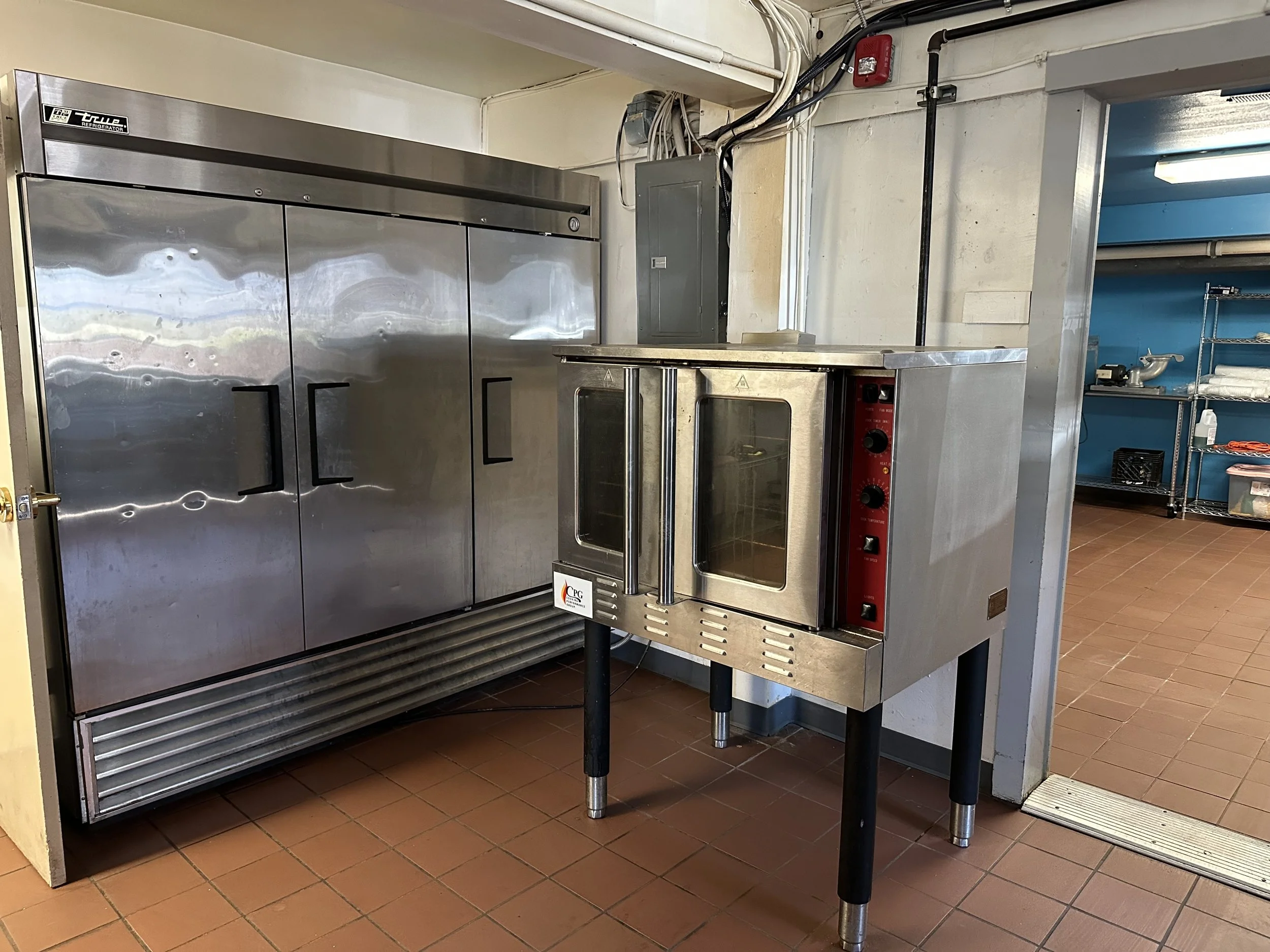A commercial kitchen scene with a large stainless steel refrigerator on the left side, and a smaller stainless steel oven or steamer with glass doors on the right side, placed on a tiled floor.