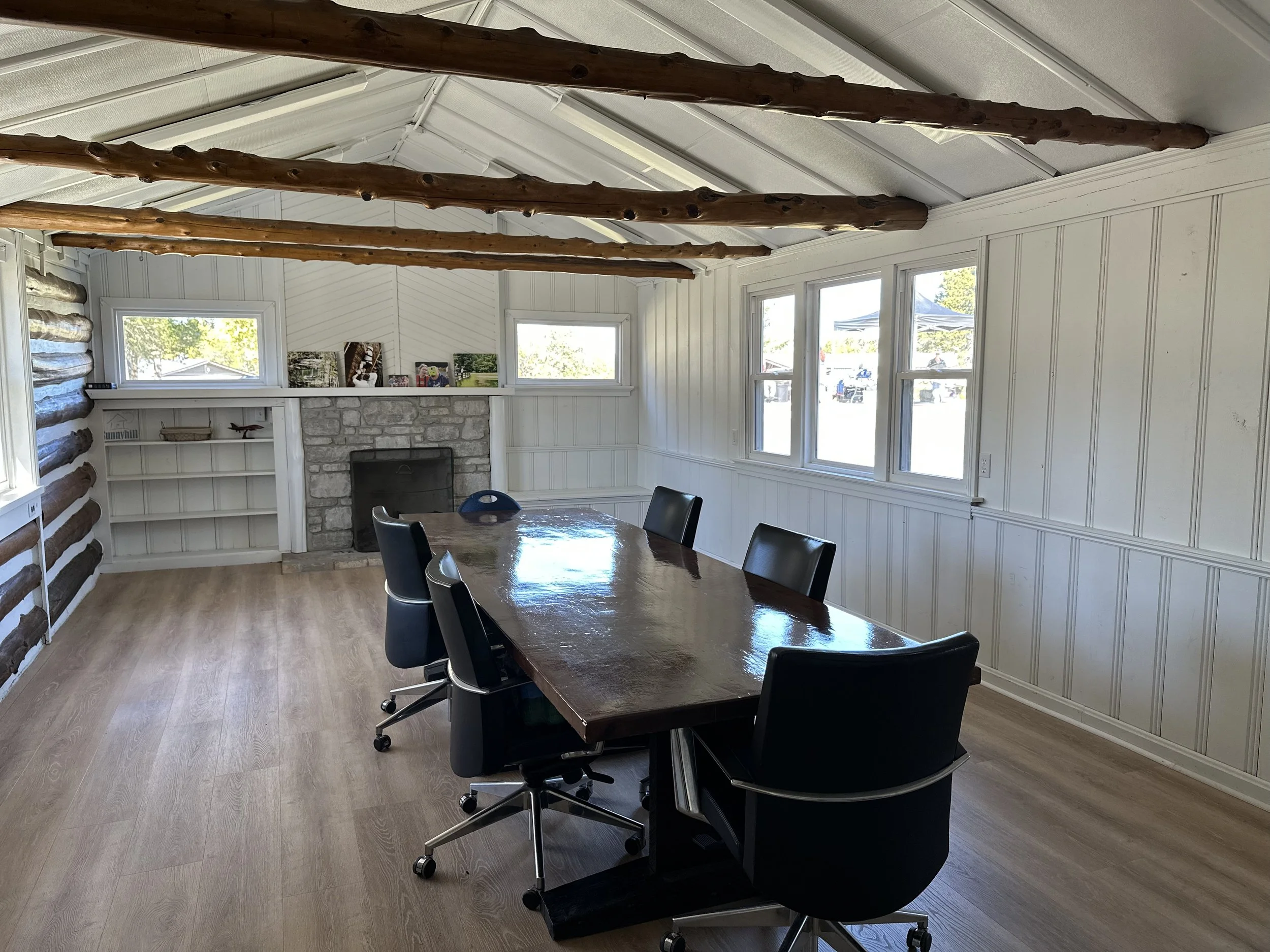 A conference room with a wooden table, six black swivel chairs, a stone fireplace, and windows providing natural light, with wood beams on the ceiling and white paneled walls.