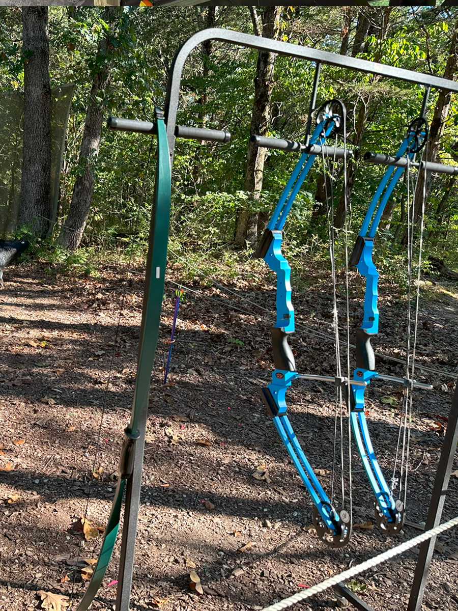 A pair of blue and black mechanical archery bows hanging under a metal frame outdoors in a wooded area with trees and dirt ground.