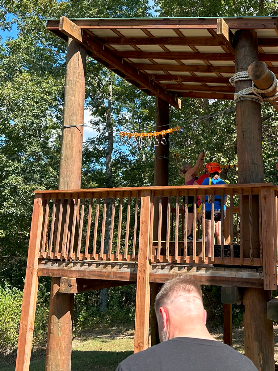 Children and an adult on a wooden treehouse with safety harnesses, in a wooded outdoor area.